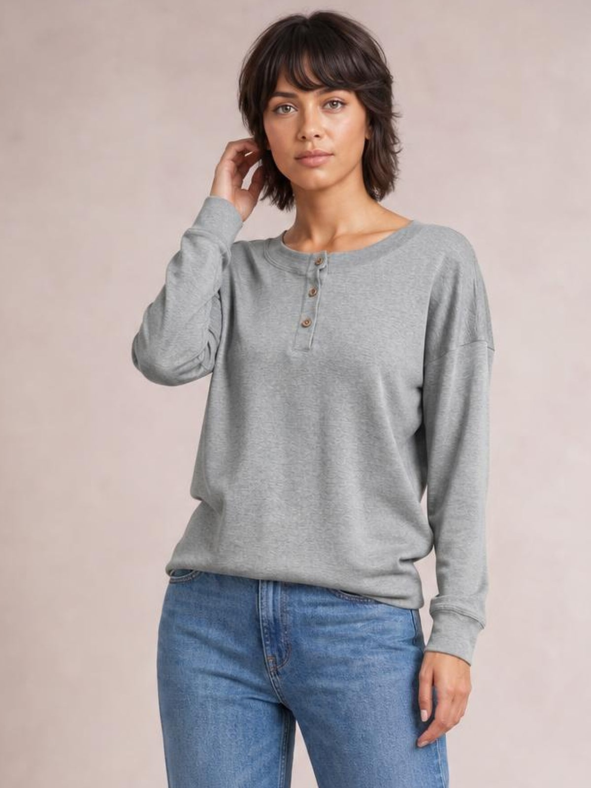Model wears a gray quarter-button dropped-shoulder blouse with a round neckline, long cuffed sleeves, and a relaxed silhouette, touching her hair.