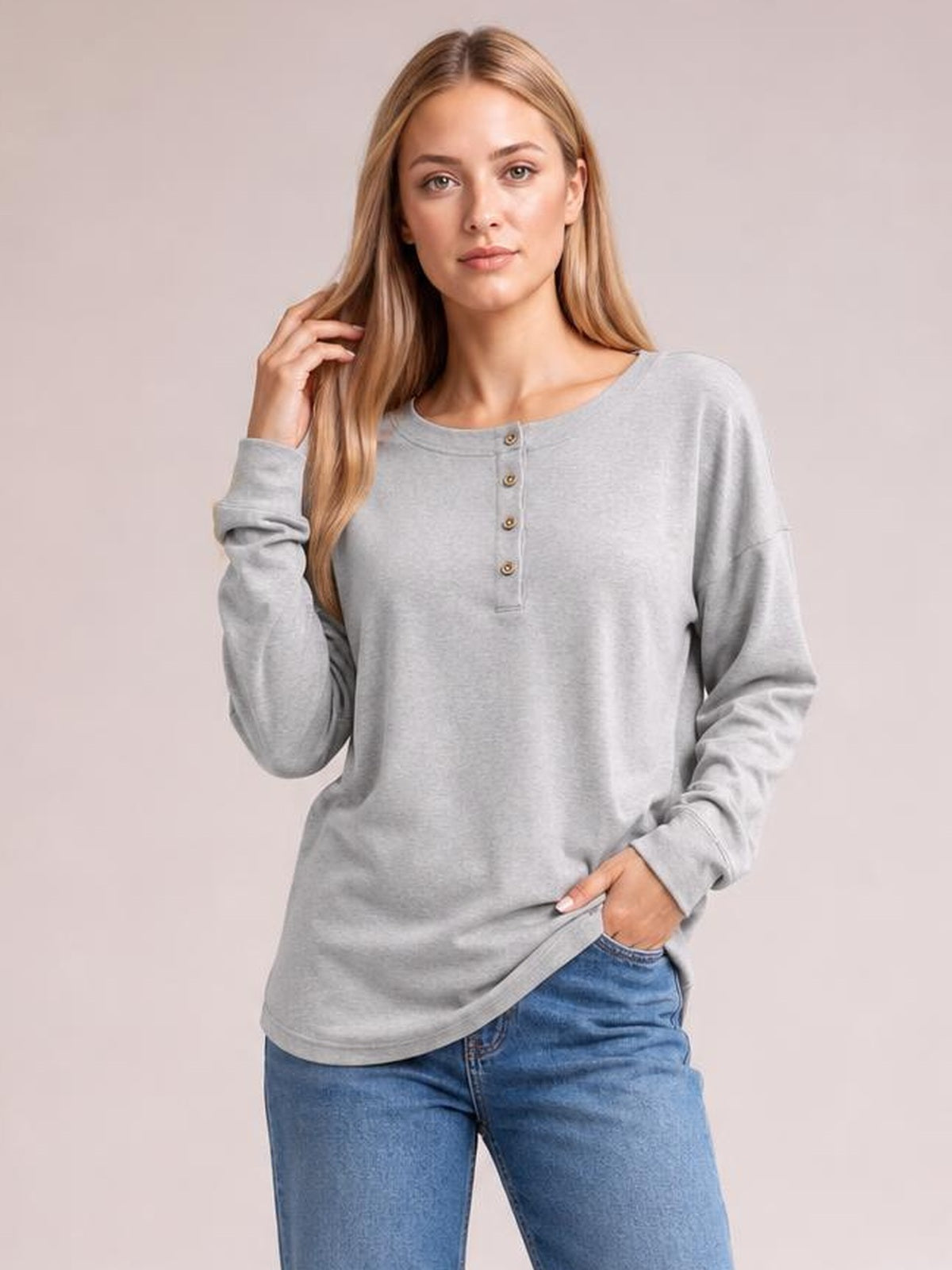 A model wears a light gray quarter-button dropped-shoulder blouse with long sleeves and a relaxed silhouette, touching her hair with one hand.