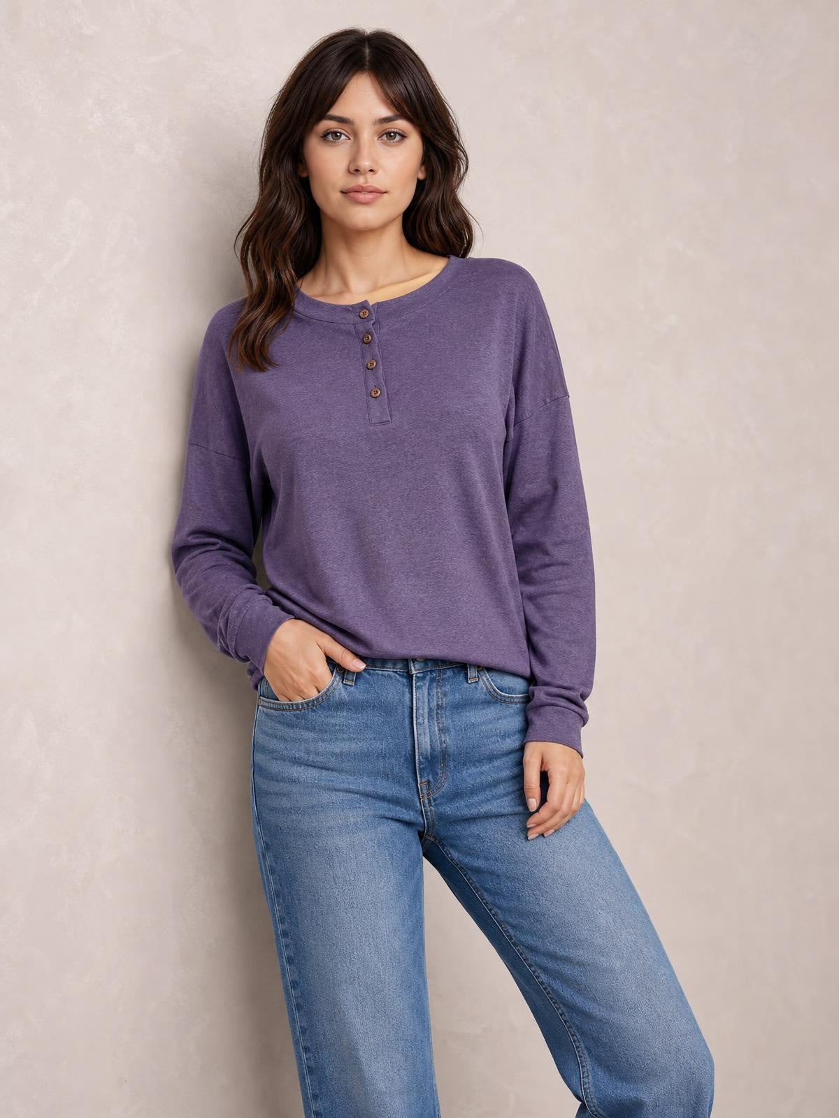 Model wearing a purple quarter-button blouse with a dropped shoulder, crew neckline, long sleeves, and relaxed silhouette, posing with one hand in her jeans pocket.