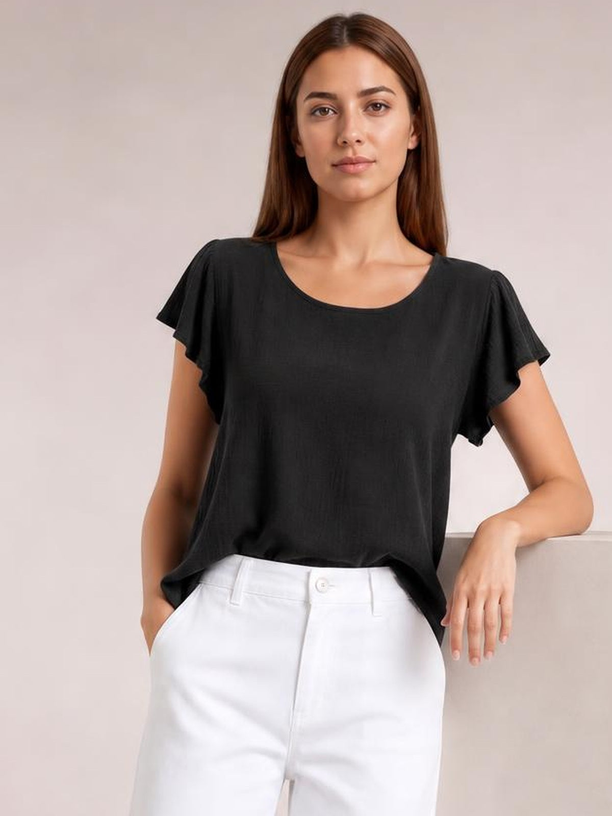 Model wears a solid black round neck top with flutter short sleeves and a relaxed regular silhouette, standing with one hand in a pocket.