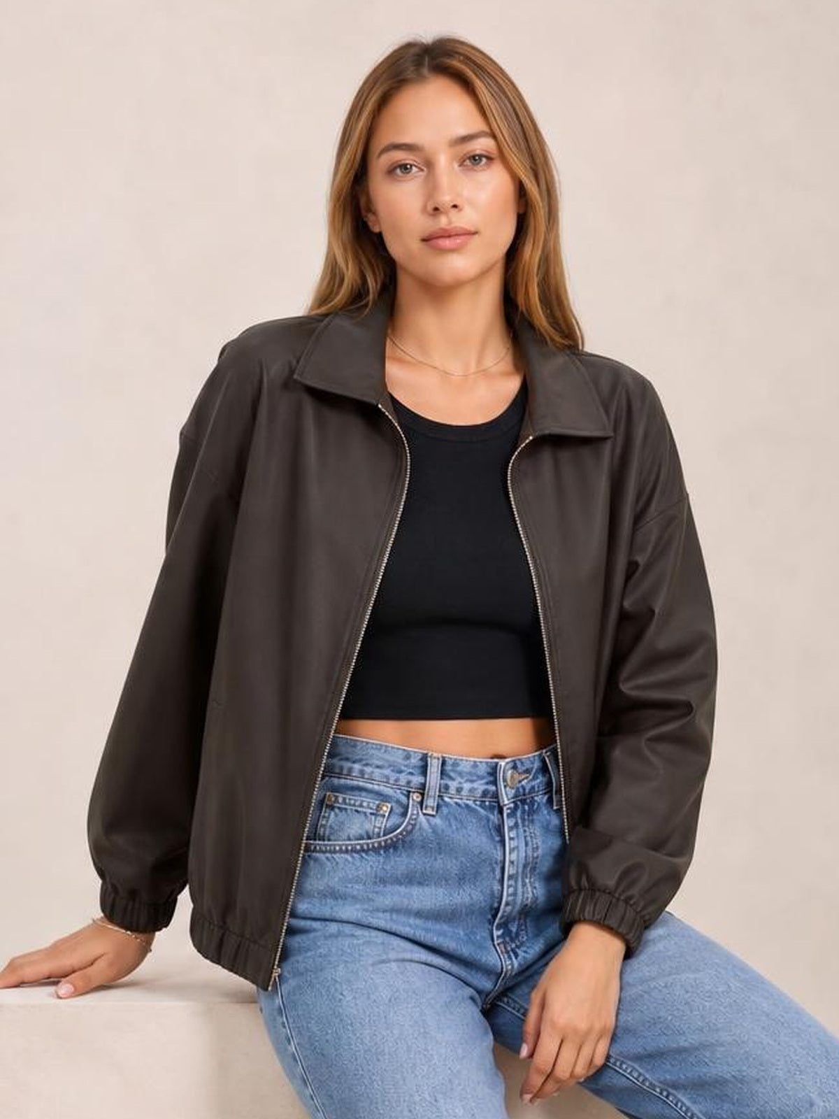 A seated model wears a dark brown zip-up dropped shoulder jacket with a relaxed silhouette, wide collar, long sleeves, and elasticized cuffs.