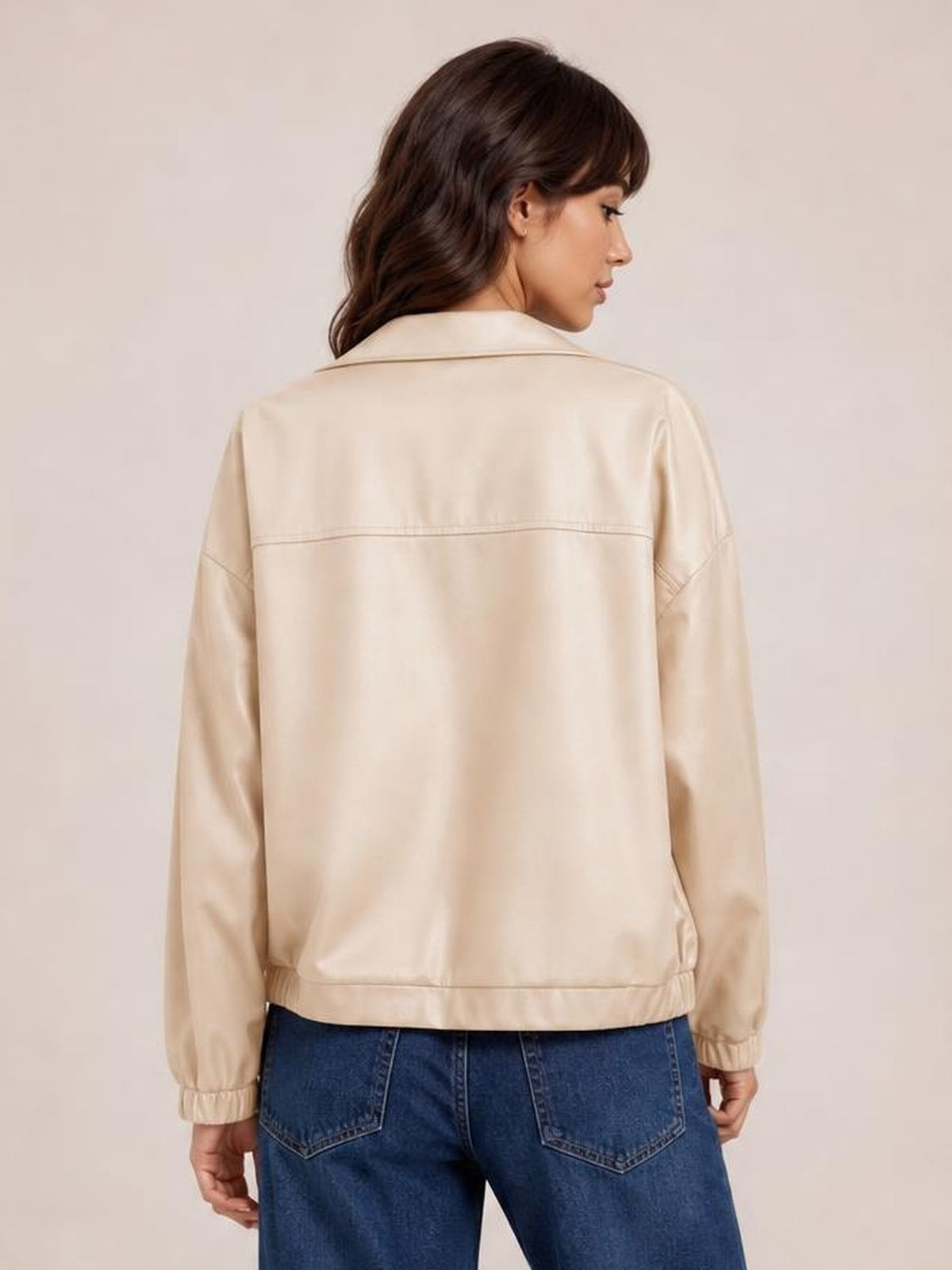 A model stands facing away in a cream zip-up dropped shoulder jacket with a relaxed silhouette, seam detailing, long sleeves, and elasticized cuffs.