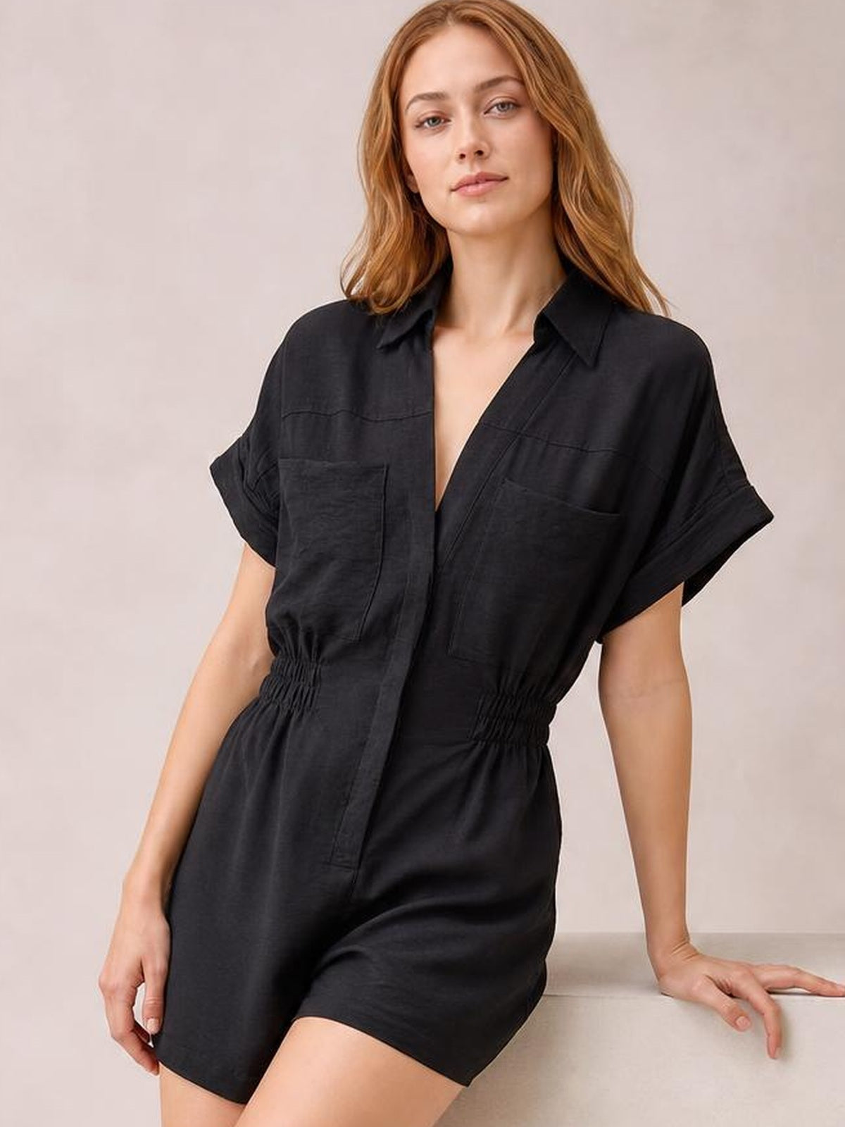 Model wearing a black Johnny collar short sleeve romper with a buttoned front, rolled cuffs, patch pockets, and a ruched elastic waist.