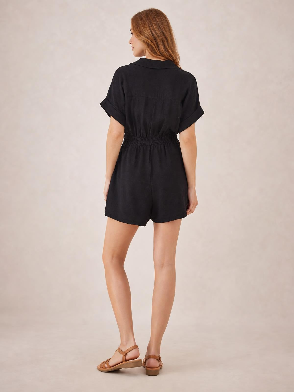 A model stands turned away in a black short-sleeve romper with a Johnny collar, rolled cuffs, relaxed shorts silhouette, and ruched elastic waist.