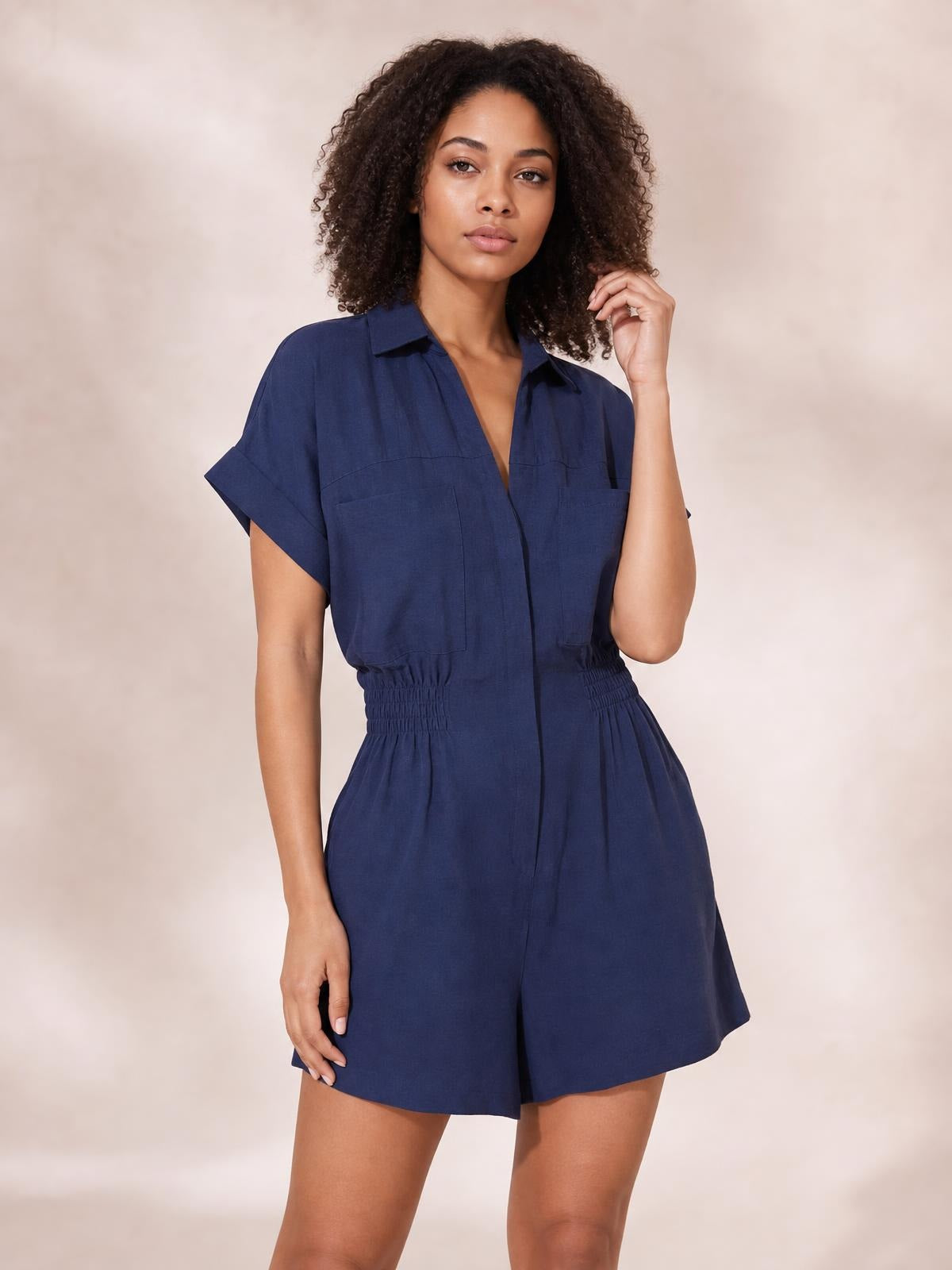 A model poses in a navy Johnny Collar short sleeve romper with a button front, chest pockets, ruched elastic waist, and relaxed shorts silhouette.