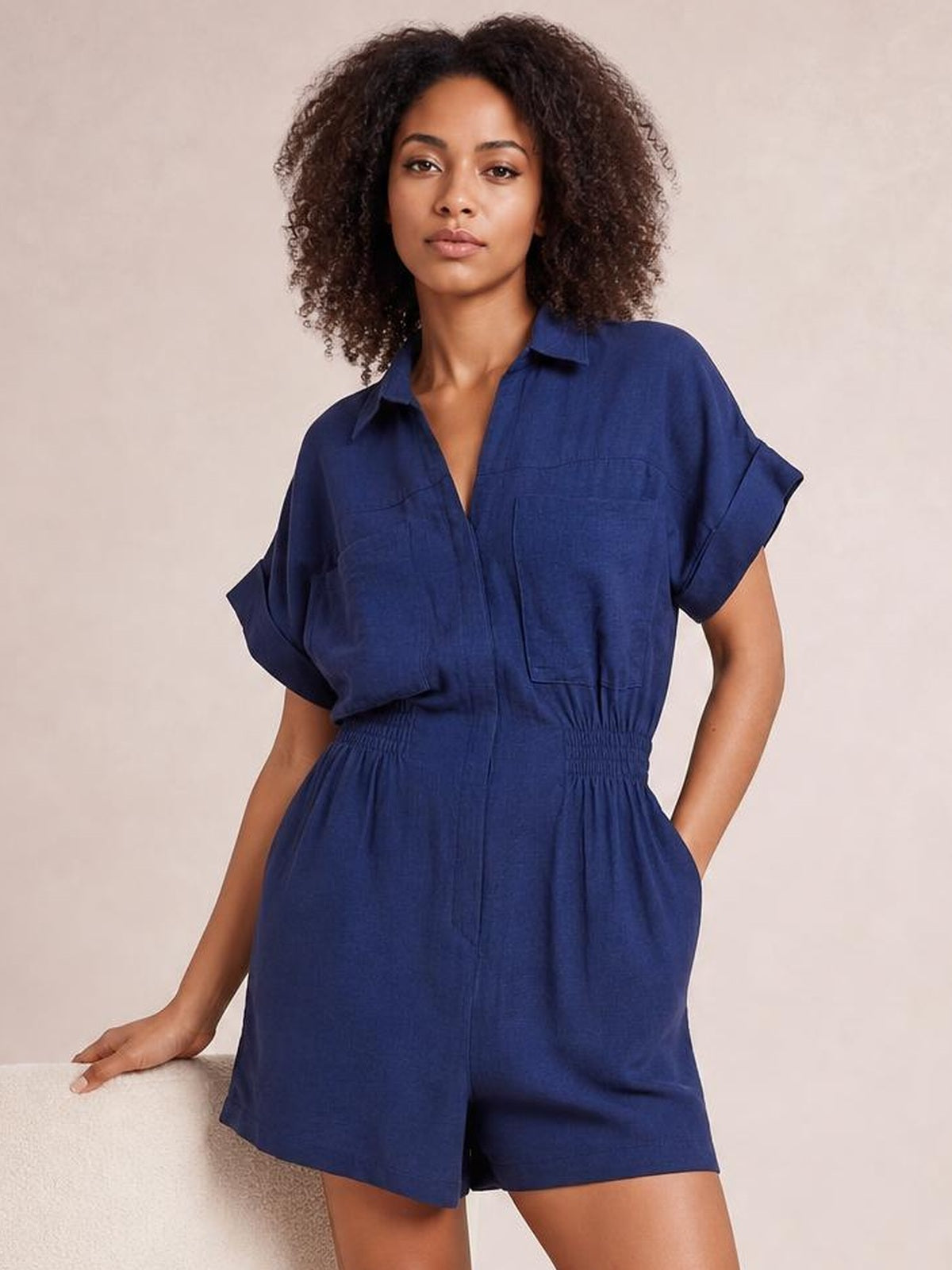 Model poses in a navy Johnny collar short sleeve romper with buttoned front, rolled cuffs, ruched waist, patch pocket, and relaxed shorts silhouette.