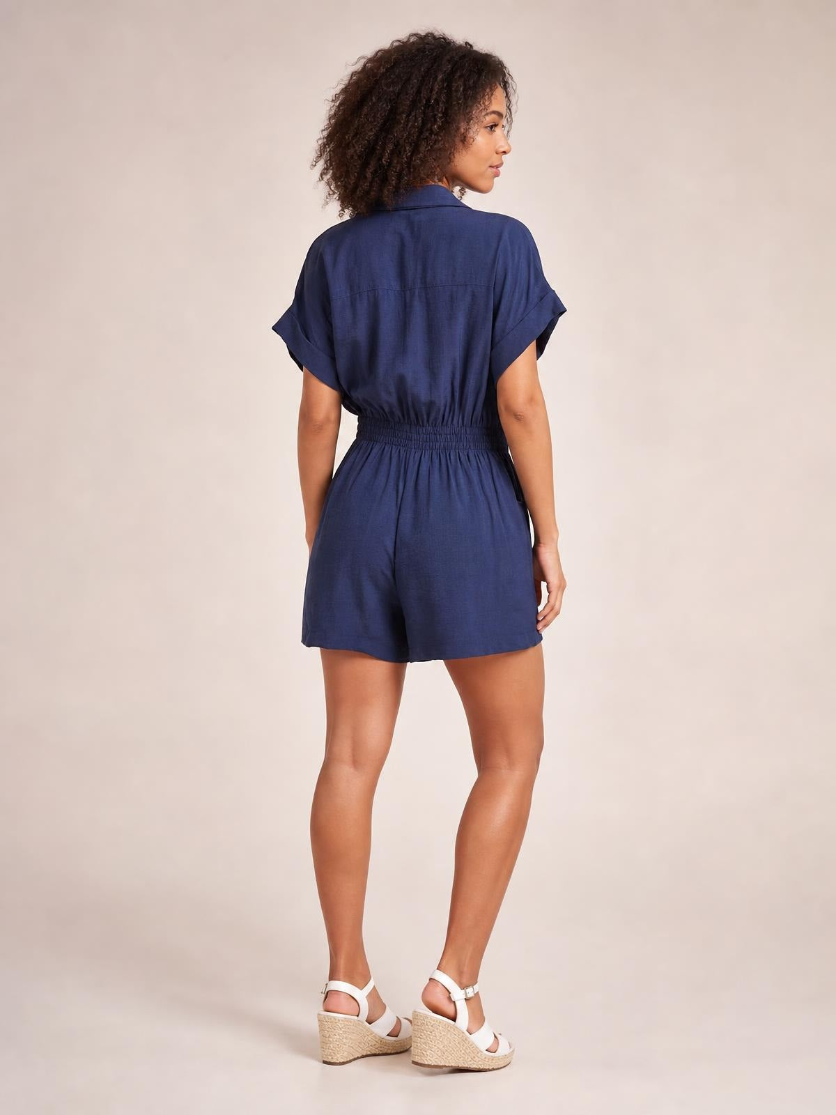 Model poses in a three-quarter back view wearing a navy Johnny Collar short sleeve romper with cuffed sleeves, ruched elastic waist, and relaxed shorts.