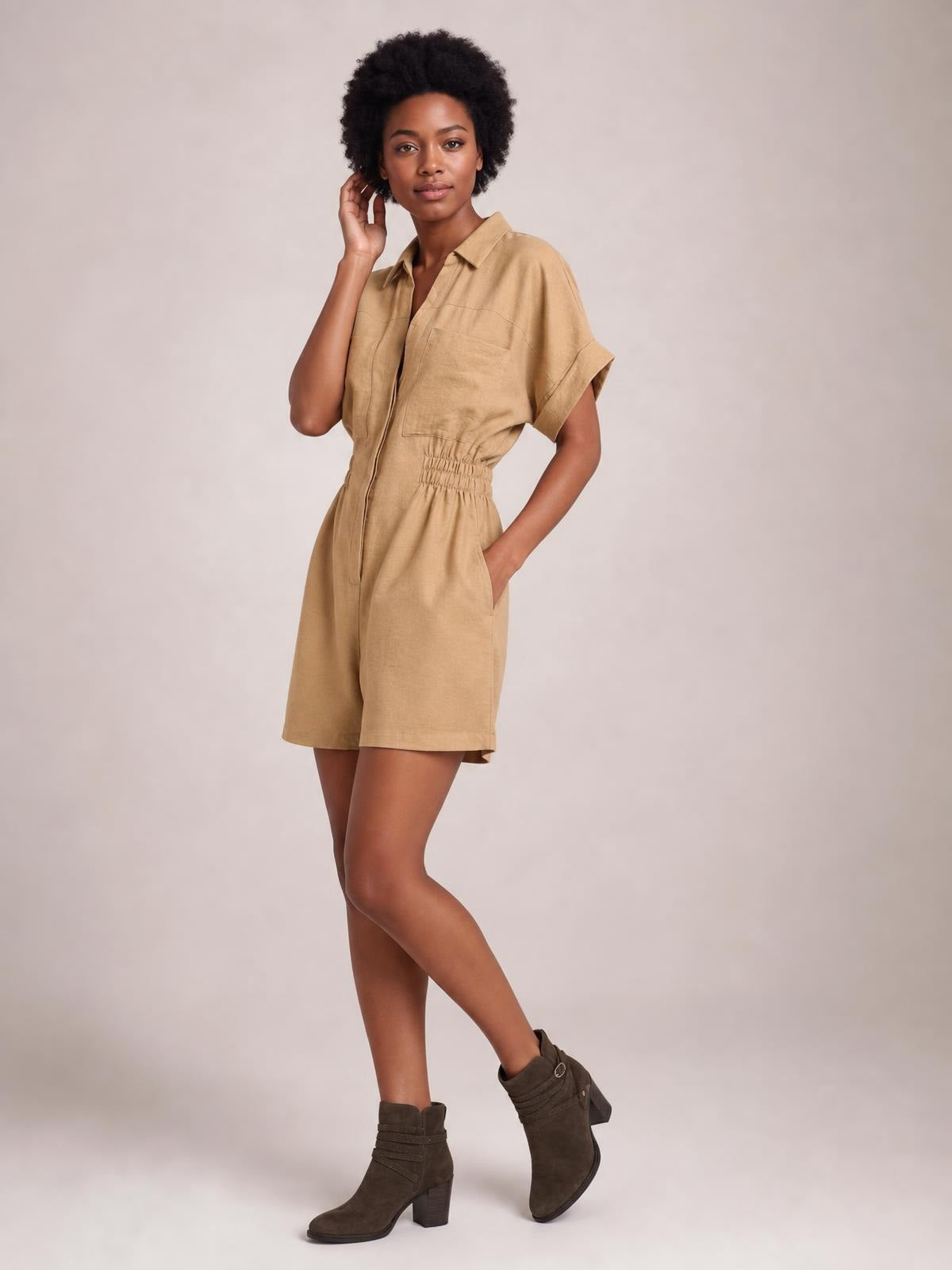 Model poses in a tan short-sleeve romper with a Johnny collar, button front, chest and side pockets, ruched elastic waist, relaxed shorts, and crossed legs.