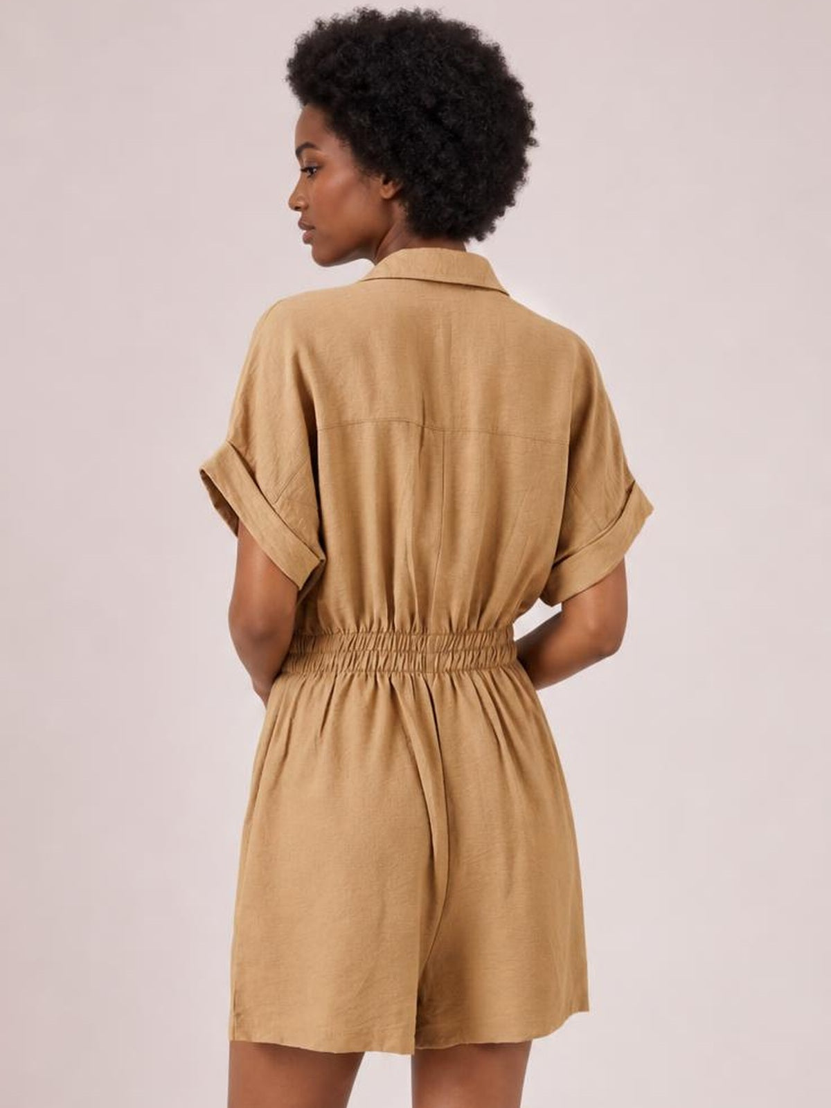A model poses from the back in a tan Johnny collar short sleeve romper with rolled cuffs, a relaxed silhouette, and a ruched elastic waist.