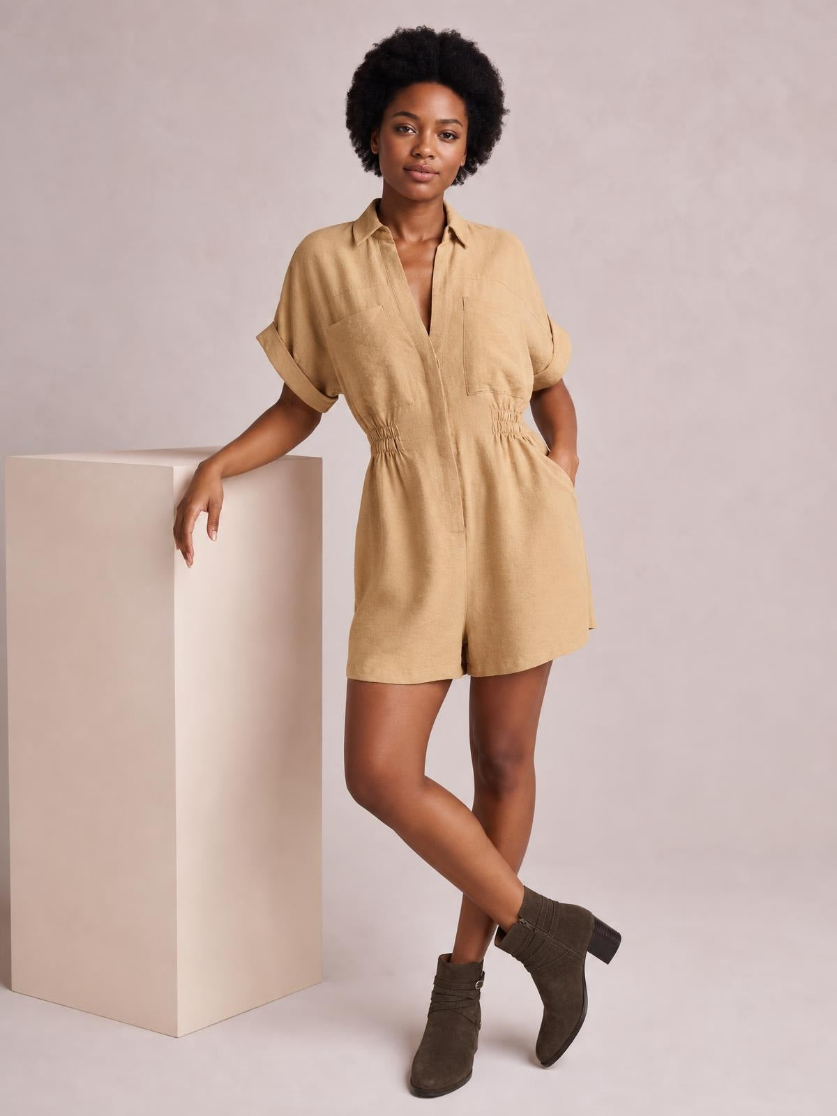 Model poses in a tan Johnny Collar short sleeve romper with a button front, rolled cuffs, ruched waist, chest pockets, and relaxed shorts silhouette.