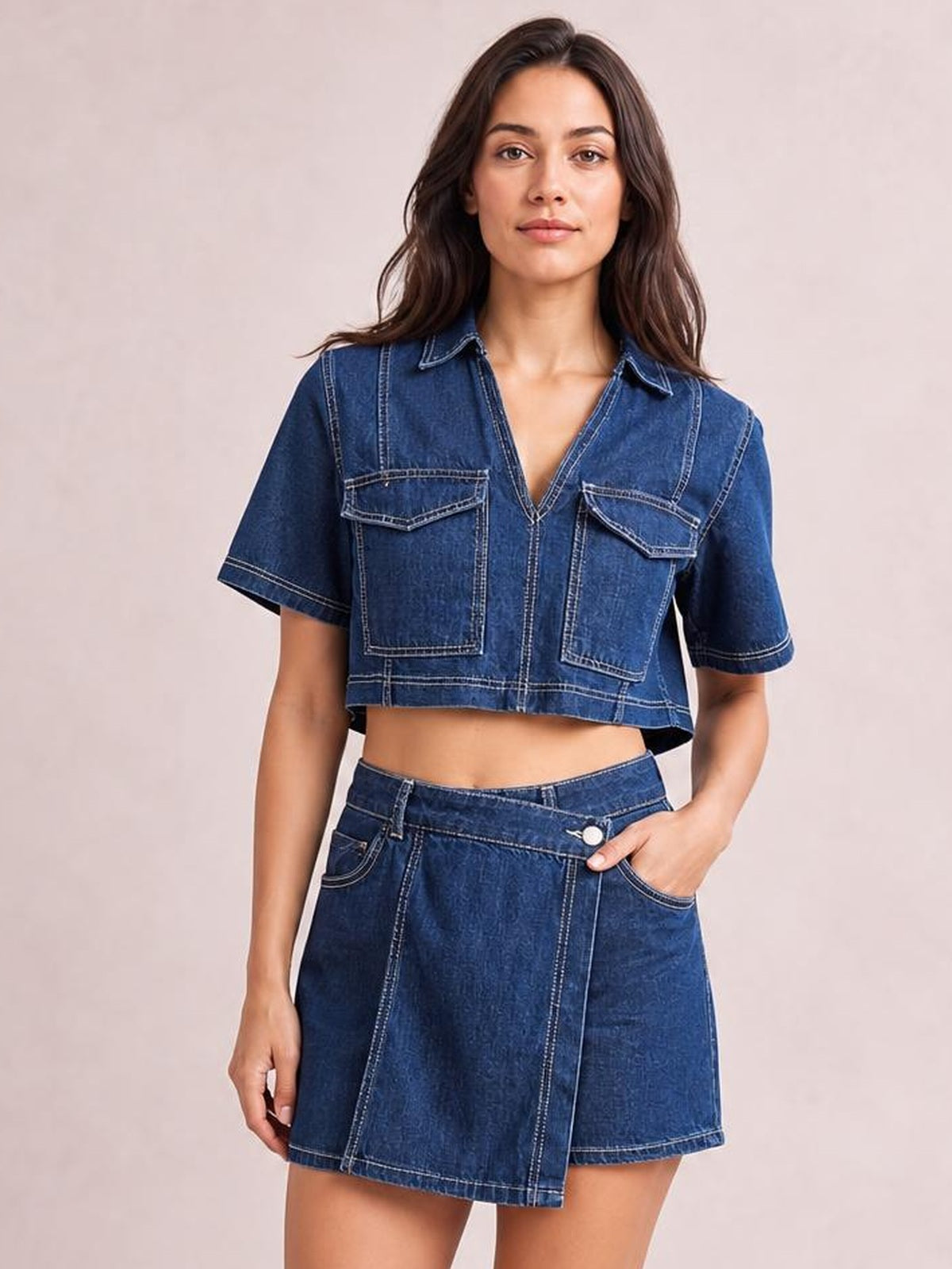 Model wears a dark blue denim set with a cropped short-sleeve Johnny collar top, contrast stitching, utility pockets, and a high-waisted wrap-front skort.