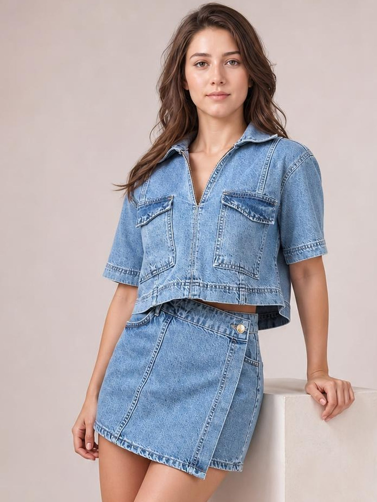 A model wears a light blue denim set with a collared short-sleeve cropped top, flap pockets, visible seams, and a structured wrap-front skort.