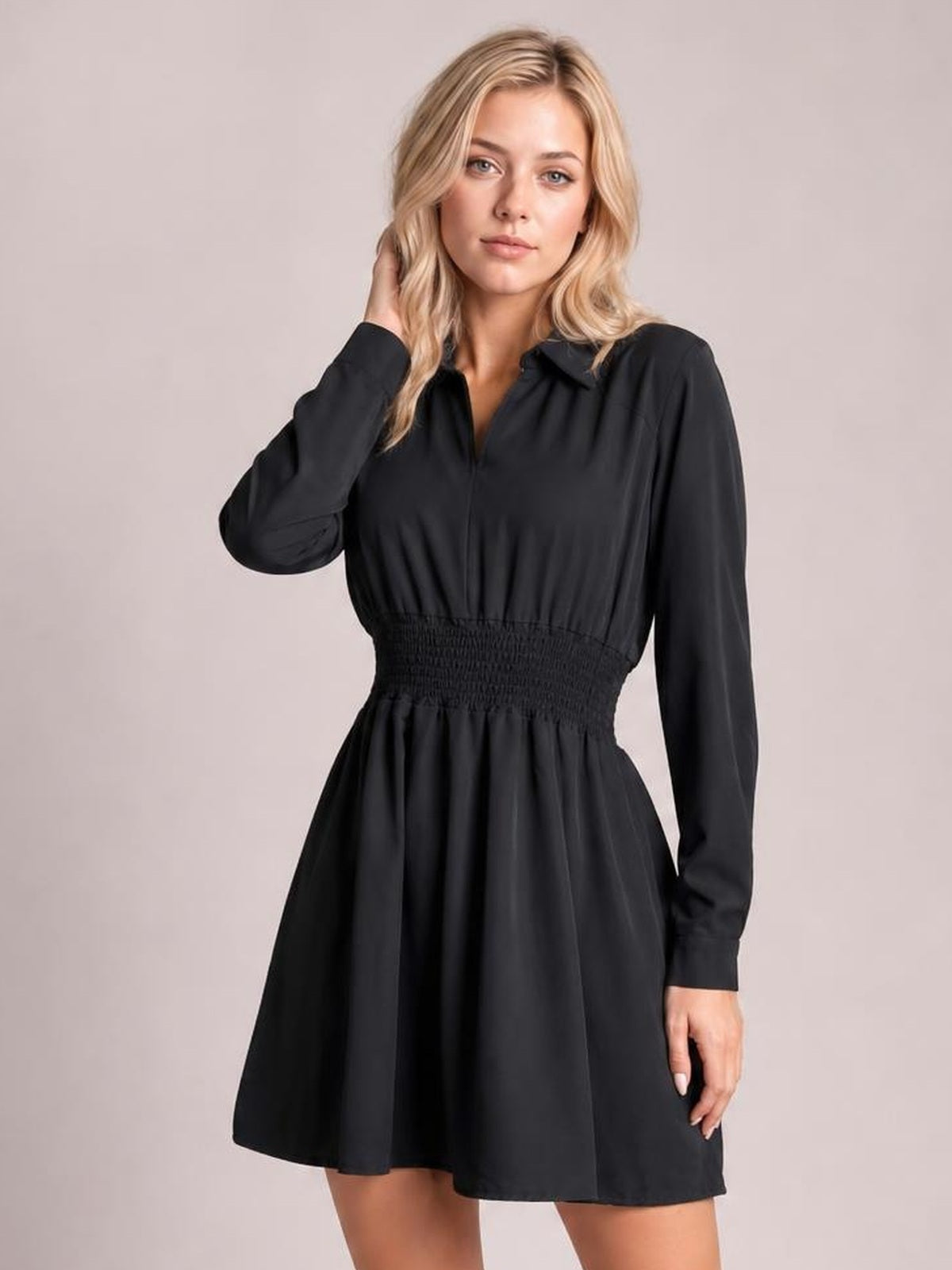 Model wearing a black Johnny collar mini dress with long sleeves, a smocked waist, and a softly flared silhouette, touching her hair.