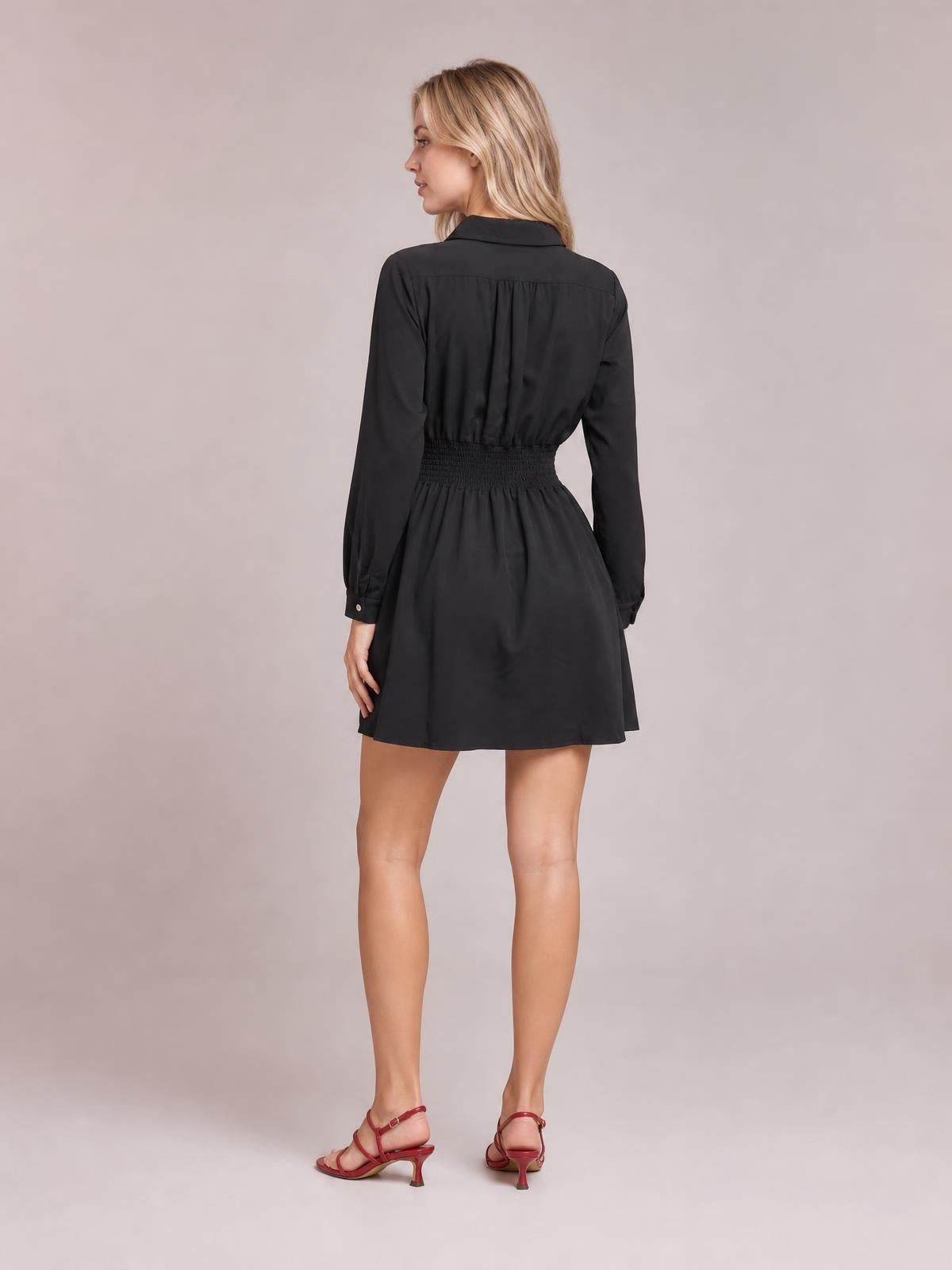 A model poses from the back in a black Johnny collar mini dress with long sleeves, a smocked waist, gathered construction, and a flared skirt.
