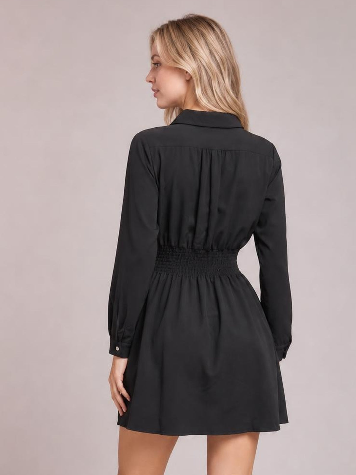 A model poses in a black long-sleeve mini dress with a Johnny collar, smocked waist, gathered back, and softly flared silhouette.