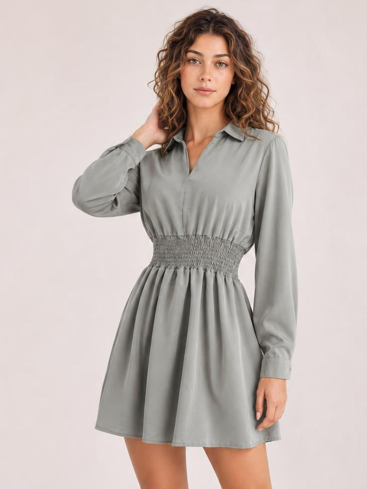A model poses in a gray Johnny collar mini dress with long sleeves, a smocked waist, and a softly flared silhouette, touching her hair.