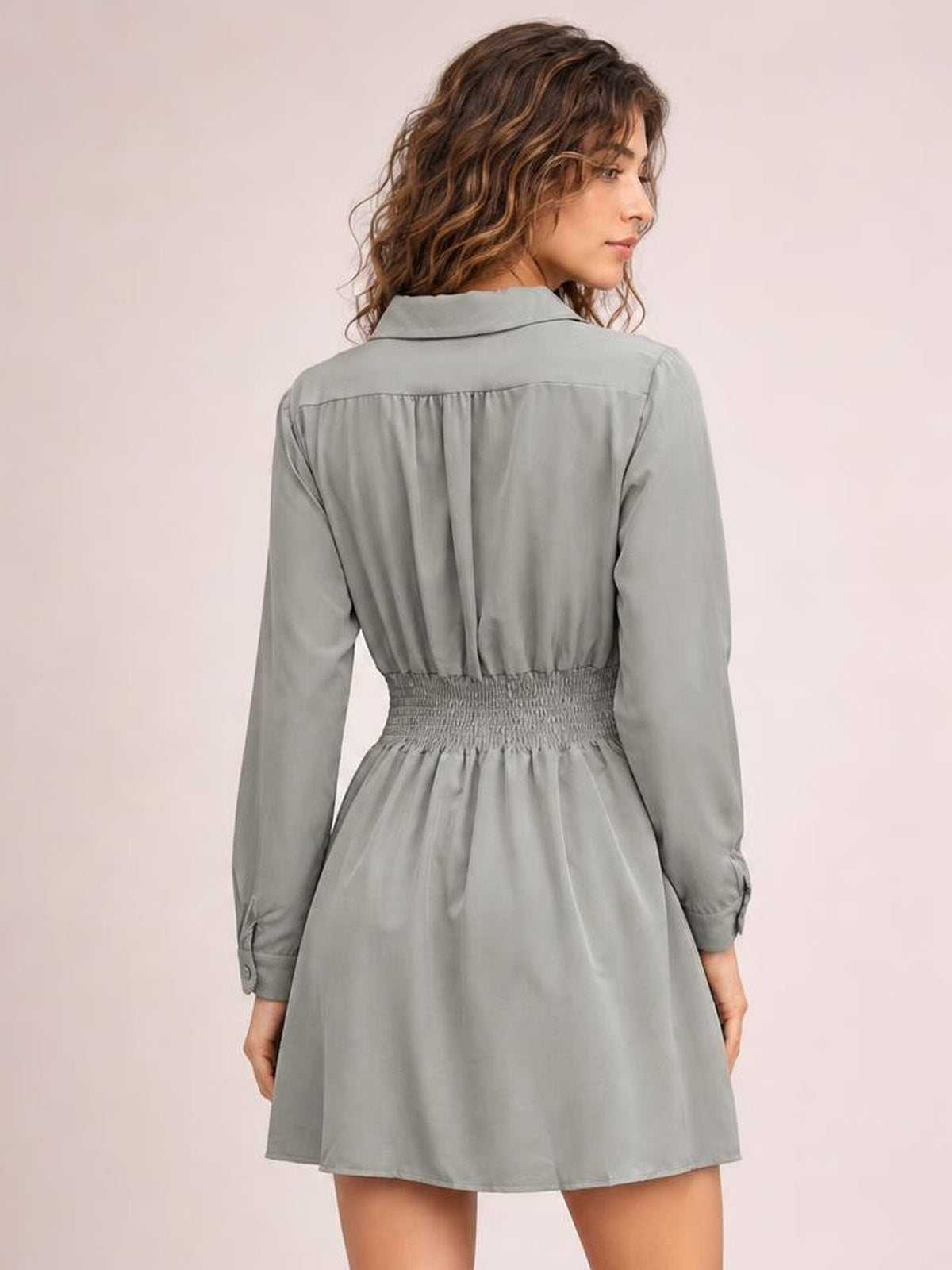 A model looks over her shoulder in a gray long-sleeve mini dress with a Johnny collar, smocked waist, gathered back, and flared silhouette.