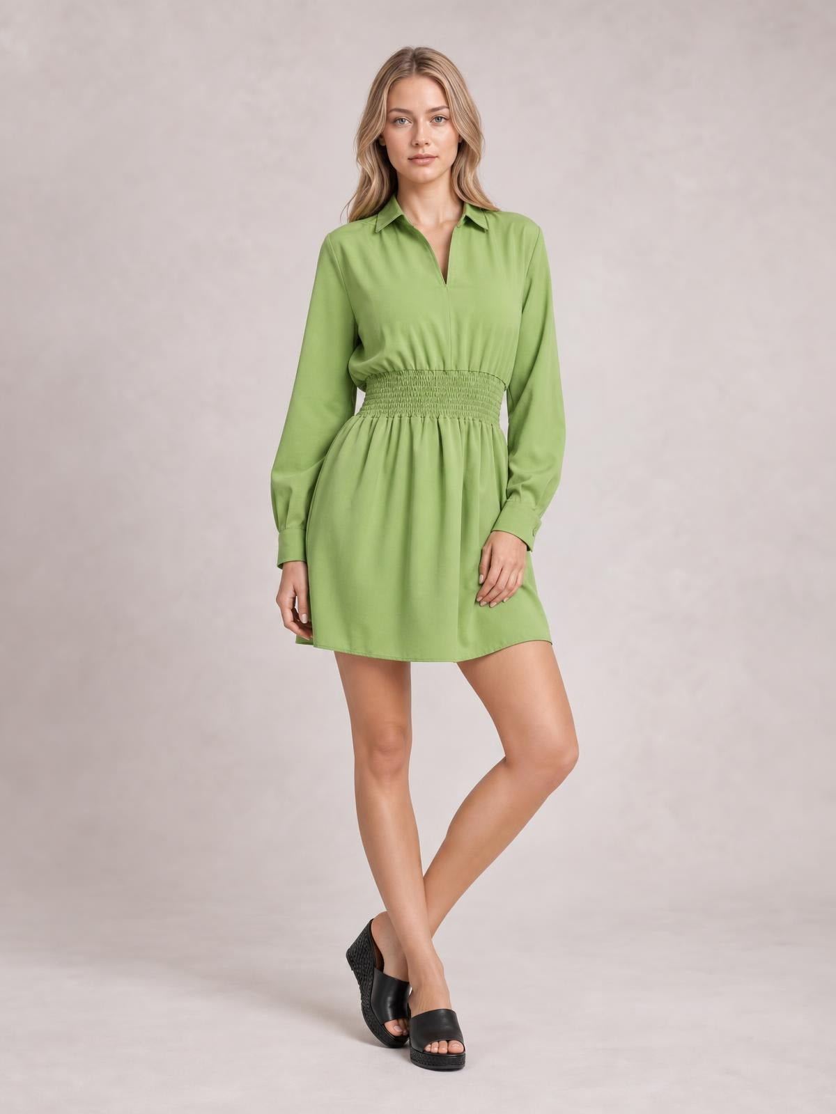 A model poses with crossed legs in a green long-sleeve mini dress featuring a Johnny collar, button-front design, smocked waist, and softly flared silhouette.