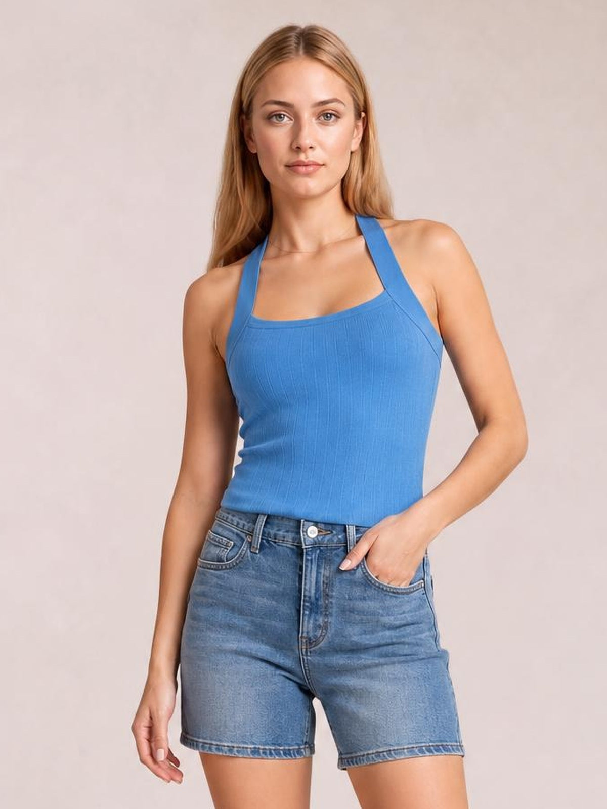 A woman modeling a bright blue ribbed halter tank top paired with blue denim shorts against a solid light beige studio background.