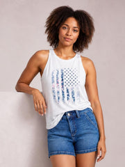 A woman wearing a white sleeveless tank top with a distressed blue American flag graphic and blue denim shorts, posing against a neutral light gray wall.