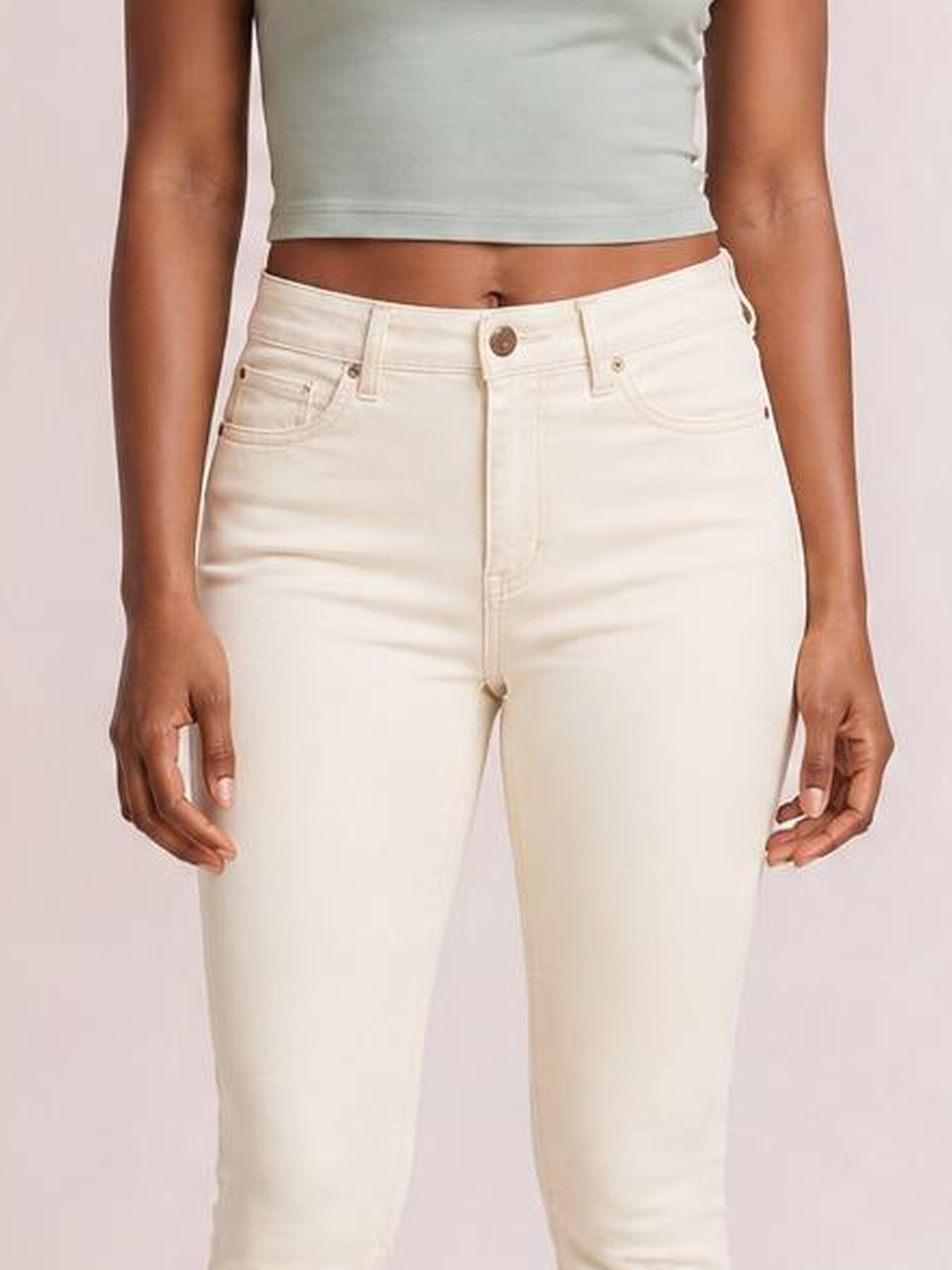 Model stands with hands at her sides wearing cream garment-dyed tummy control skinny jeans with a high-rise waist, belt loops, button fly, and five-pocket construction.