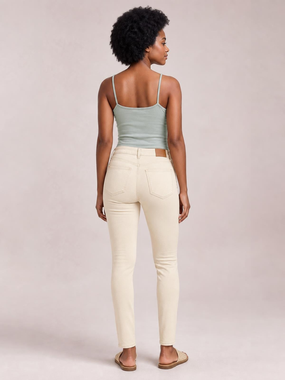 A model stands in a rear view wearing light cream garment-dyed tummy control skinny jeans with a high-rise waistband, belt loops, and back pockets.