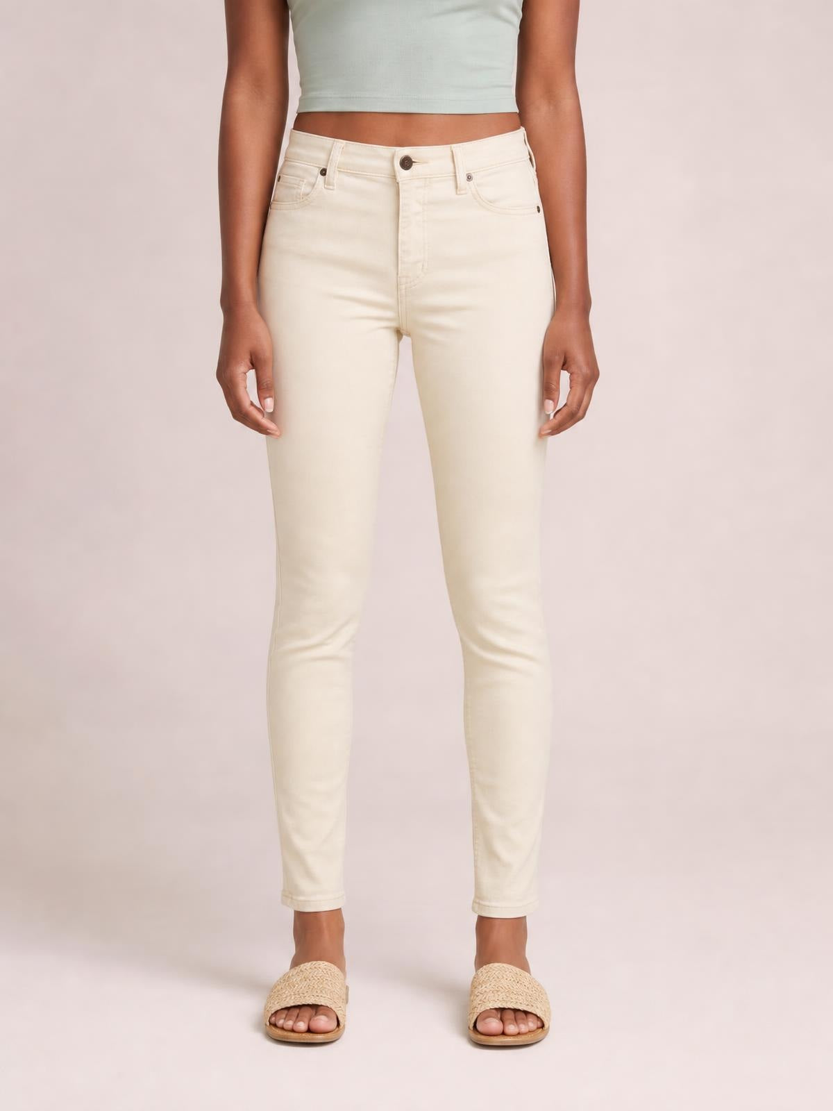 Model stands with hands at her sides wearing cream garment-dyed tummy control skinny jeans with a high-rise waist, front pockets, button closure, and slim ankle-length silhouette.