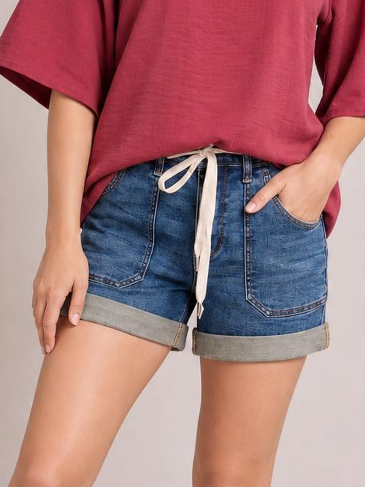 Model poses in dark blue high-rise cuff denim shorts with large front pockets, belt loops, a drawstring waist, and wide rolled hems.