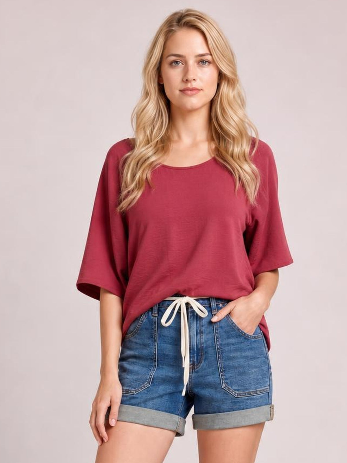 A woman modeling a loose-fitting, short-sleeve scoop-neck top in a dusty rose or burgundy shade, paired with blue denim shorts with a white drawstring, against a solid light gray studio background.