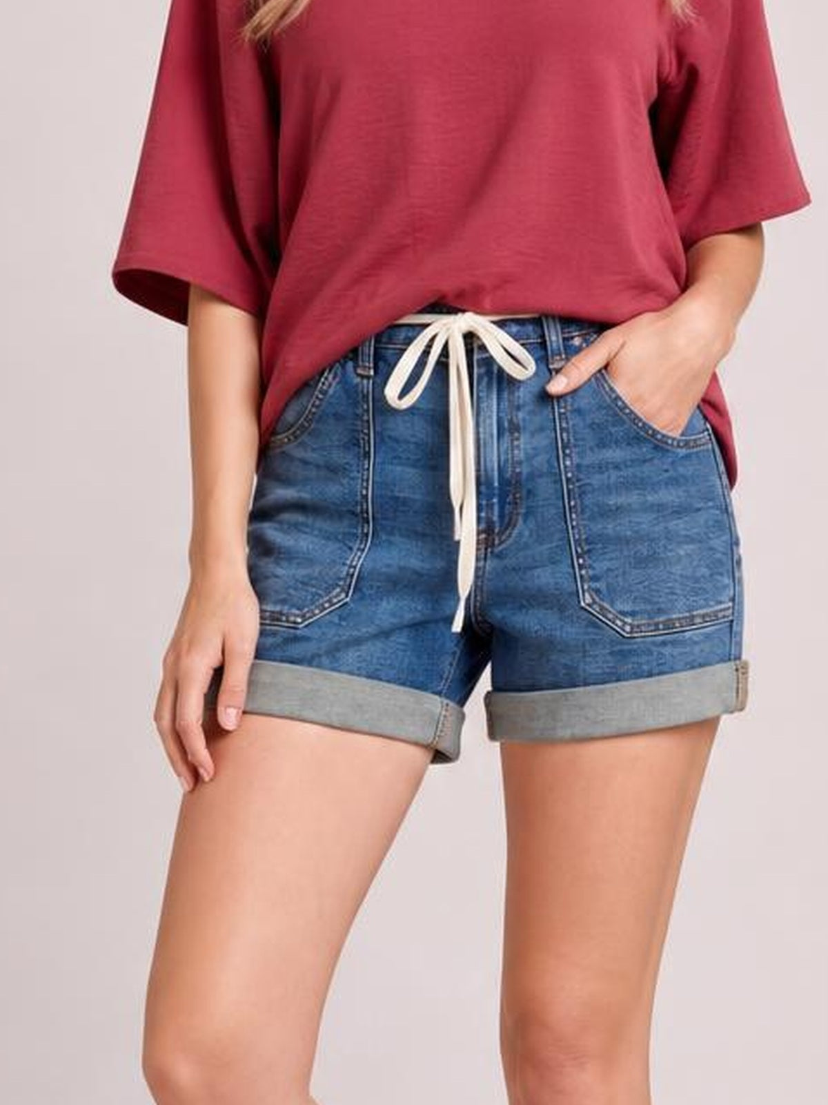 Model wears high-rise blue denim shorts with large front pockets, a drawstring waist, and wide rolled cuffs in a relaxed fit.