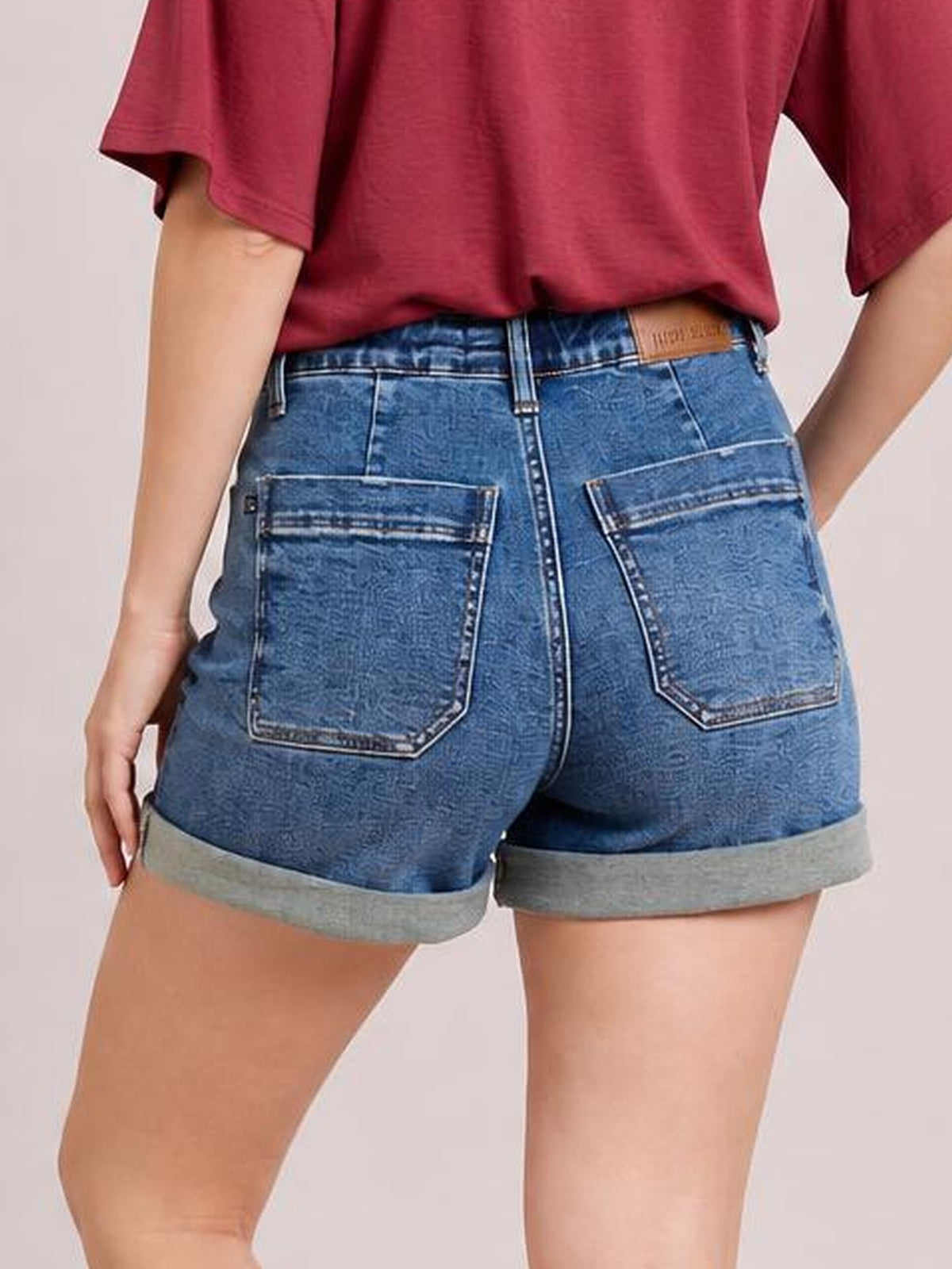 Model shown from the back wearing medium-wash high-rise cuff denim shorts with a fitted silhouette, rolled hems, belt loops, and large patch pockets.