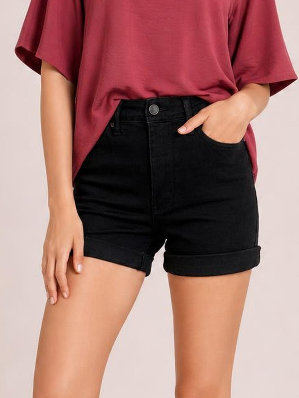 Model wearing black high-waist tummy control denim shorts with rolled cuffed hems, front button closure, and a fitted silhouette, posing with one hand in a pocket.