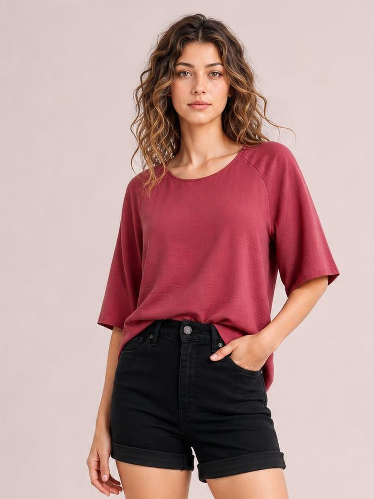 A woman modeling a loose-fitting, short-sleeve crewneck top in a dusty rose or mauve shade, paired with black denim shorts, against a solid pale pink studio background.