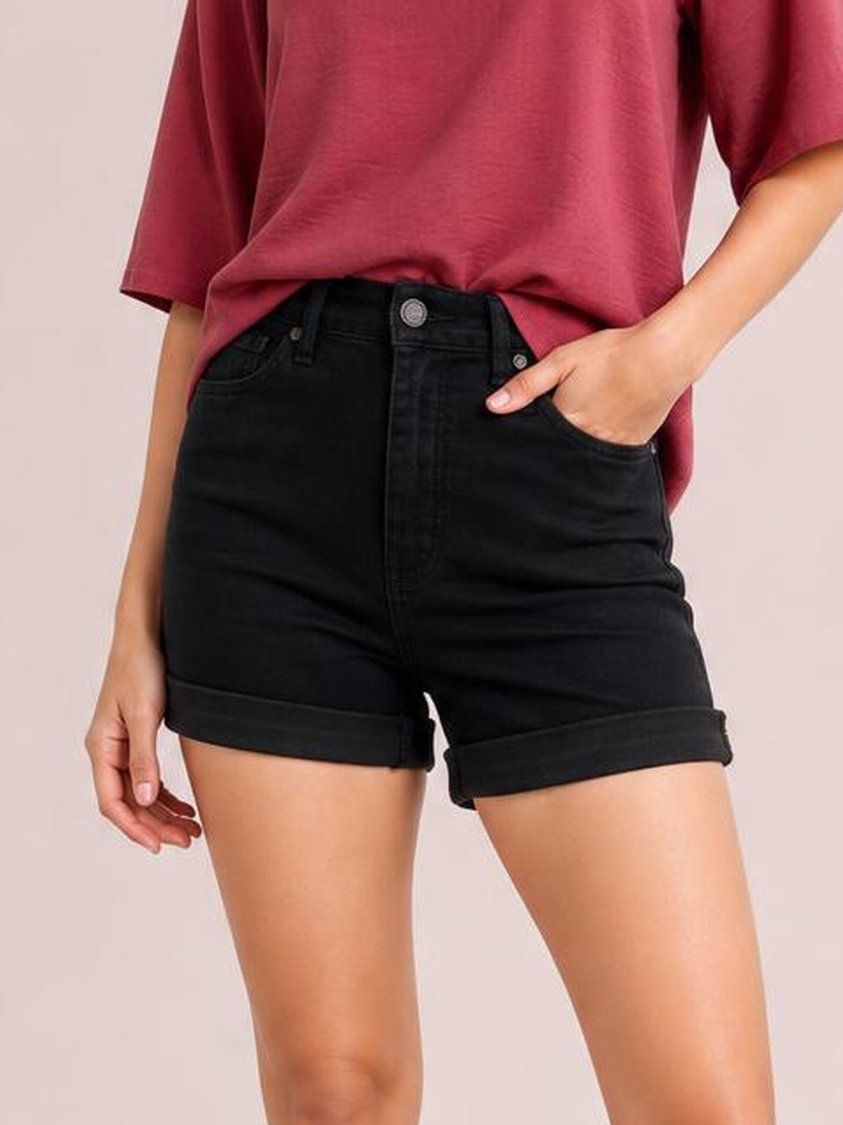 Model wears black high-waist tummy control denim shorts with cuffed hems, classic five-pocket construction, and a fitted silhouette while standing with one hand in a pocket.