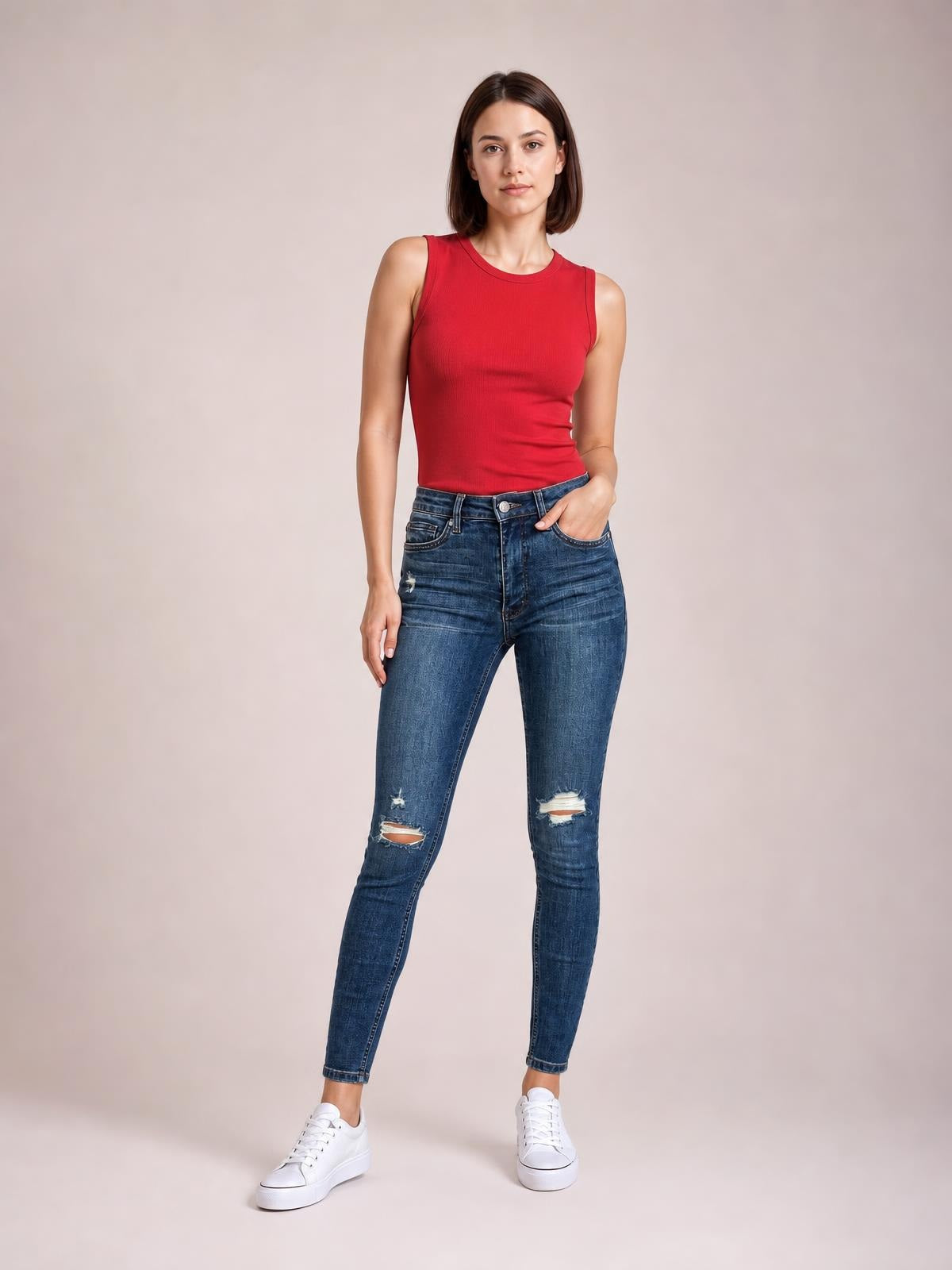 Model wearing dark blue mid-waist distressed slim jeans with front pockets, faded detailing, ripped knees, and a tapered ankle-length silhouette.