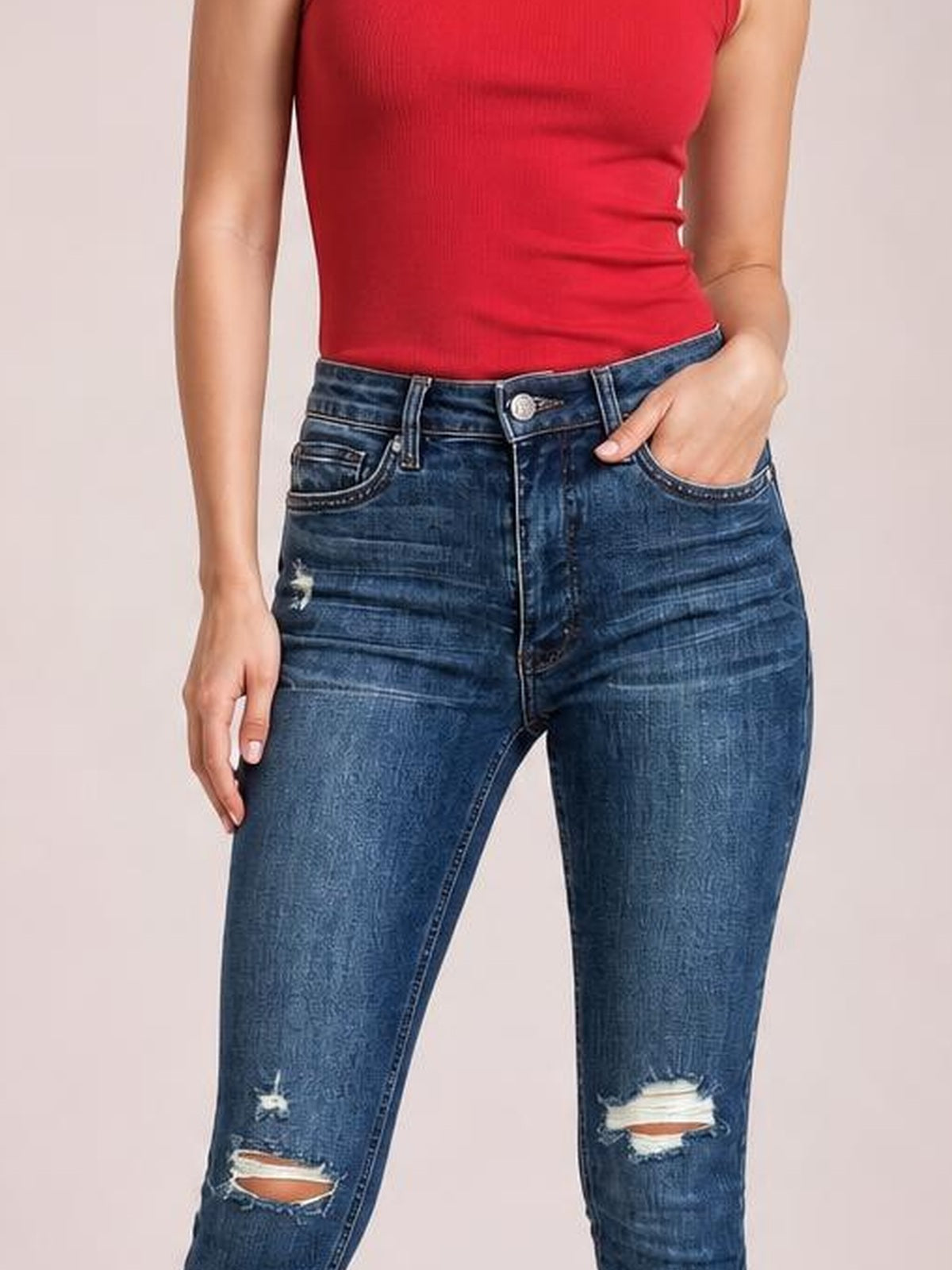 Model wearing dark blue mid-waist distressed slim jeans with front pockets, belt loops, zip fly, and ripped knees, standing with one hand in a pocket.