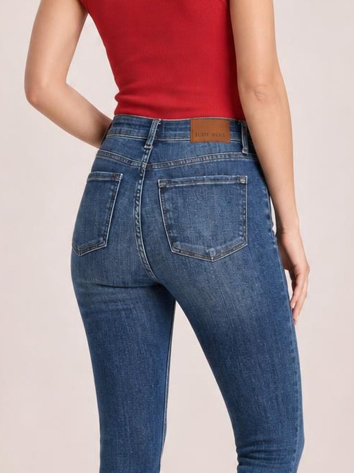 A model poses with one hand on her hip, showing the back of dark blue mid-waist slim jeans with belt loops, contrast stitching, and patch pockets.