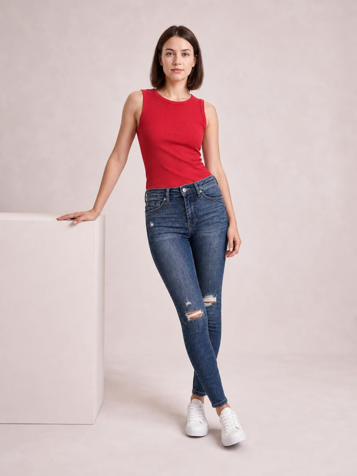 Model wearing mid-waist distressed slim jeans in dark blue denim with belt loops, pockets, a zip fly, and ripped knees, posing with crossed legs.