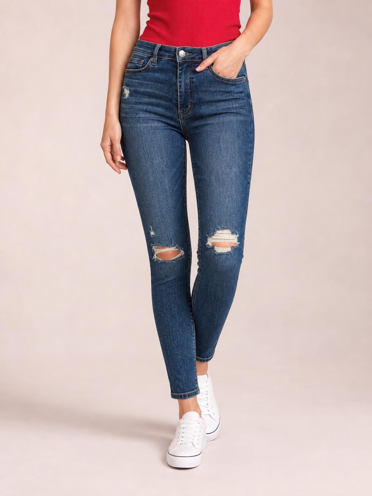 Model wearing dark blue mid-waist distressed slim jeans with five-pocket construction, ripped knees, tapered legs, and one hand tucked into a pocket.