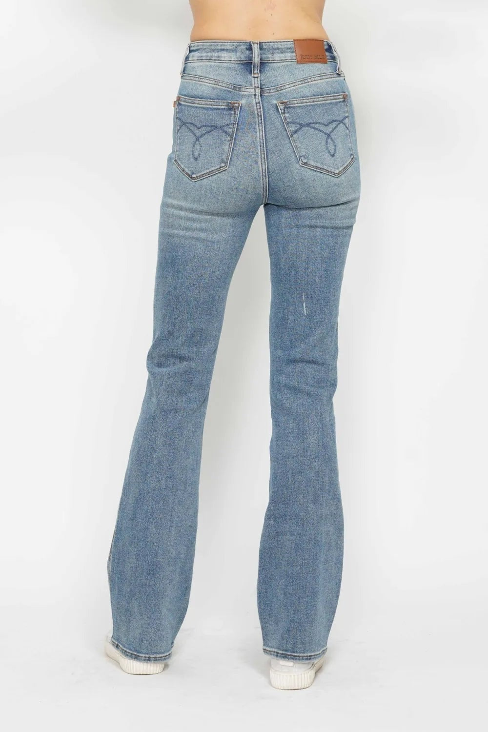 Model stands in a rear view wearing light-wash tummy control bootcut jeans with embroidered back pockets, belt loops, and a fitted stretch silhouette.