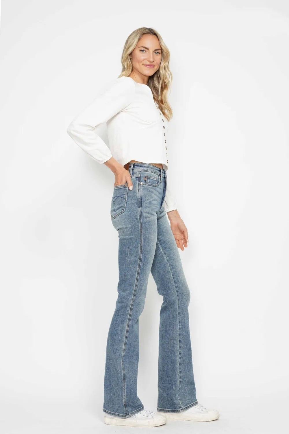 Model poses with one hand in the pocket, showcasing light-wash tummy control jeans with a high-rise waist, embroidered back pocket, and fitted bootcut legs.