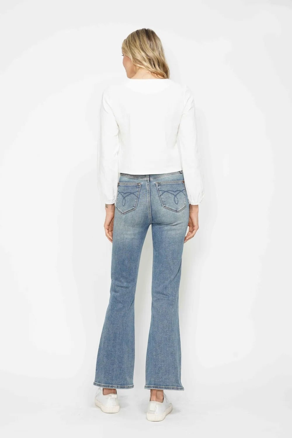 Model shown from the back in light-wash tummy control bootcut jeans with embroidered back pockets, a fitted silhouette, and flared hems.