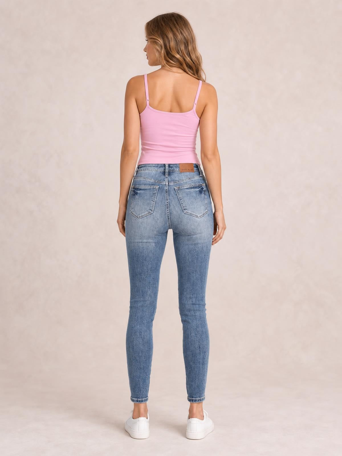 A model stands facing away in high-rise JuBlue tummy control skinny jeans with a light blue contrast wash, fitted silhouette, waistband, and back pockets.