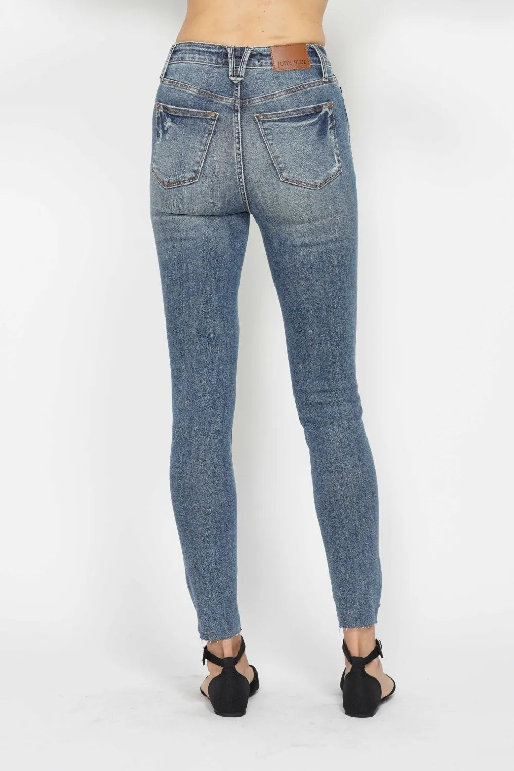 Back view of a model wearing blue vintage-wash tummy control skinny jeans with belt loops, contrast stitching, back pockets, and distressed raw hems.