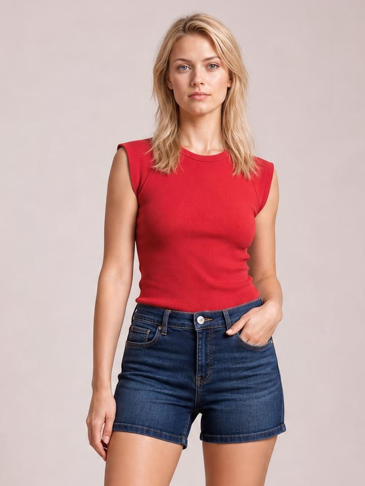 A woman modeling a sleeveless red top and dark blue denim shorts against a solid light beige studio background.
