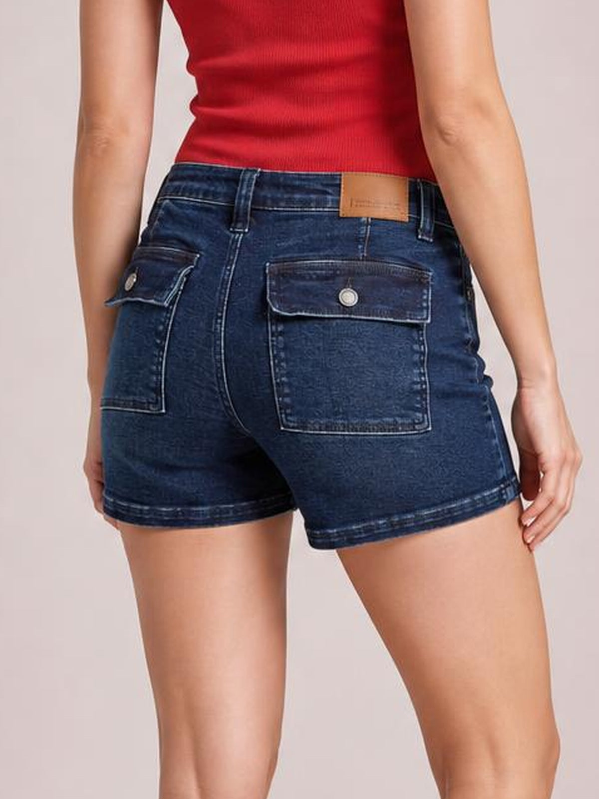 A model poses from the back wearing dark blue high-waist denim shorts with a fitted silhouette, belt loops, contrast stitching, and flap back pockets.