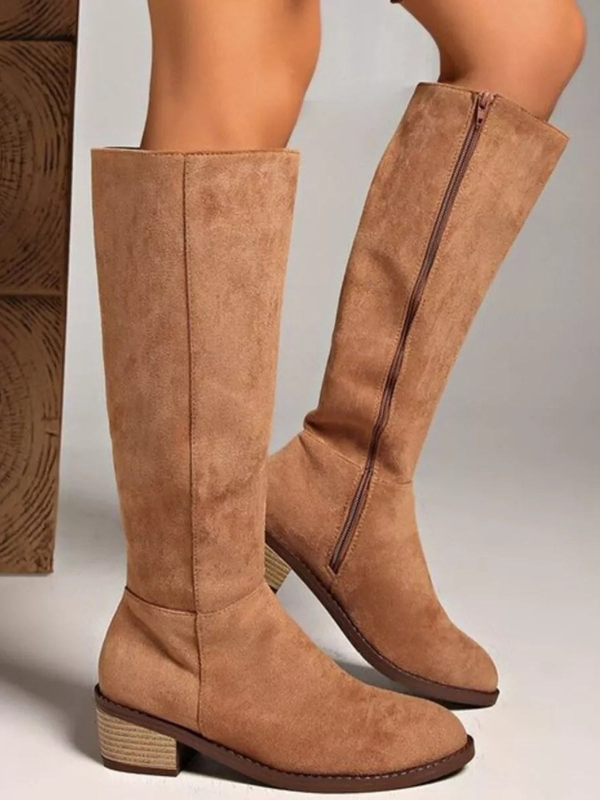 A model poses in tan suede knee-high boots with structured shafts, side zippers, rounded toes, and mid-height stacked block heels, one foot crossed behind the other.
