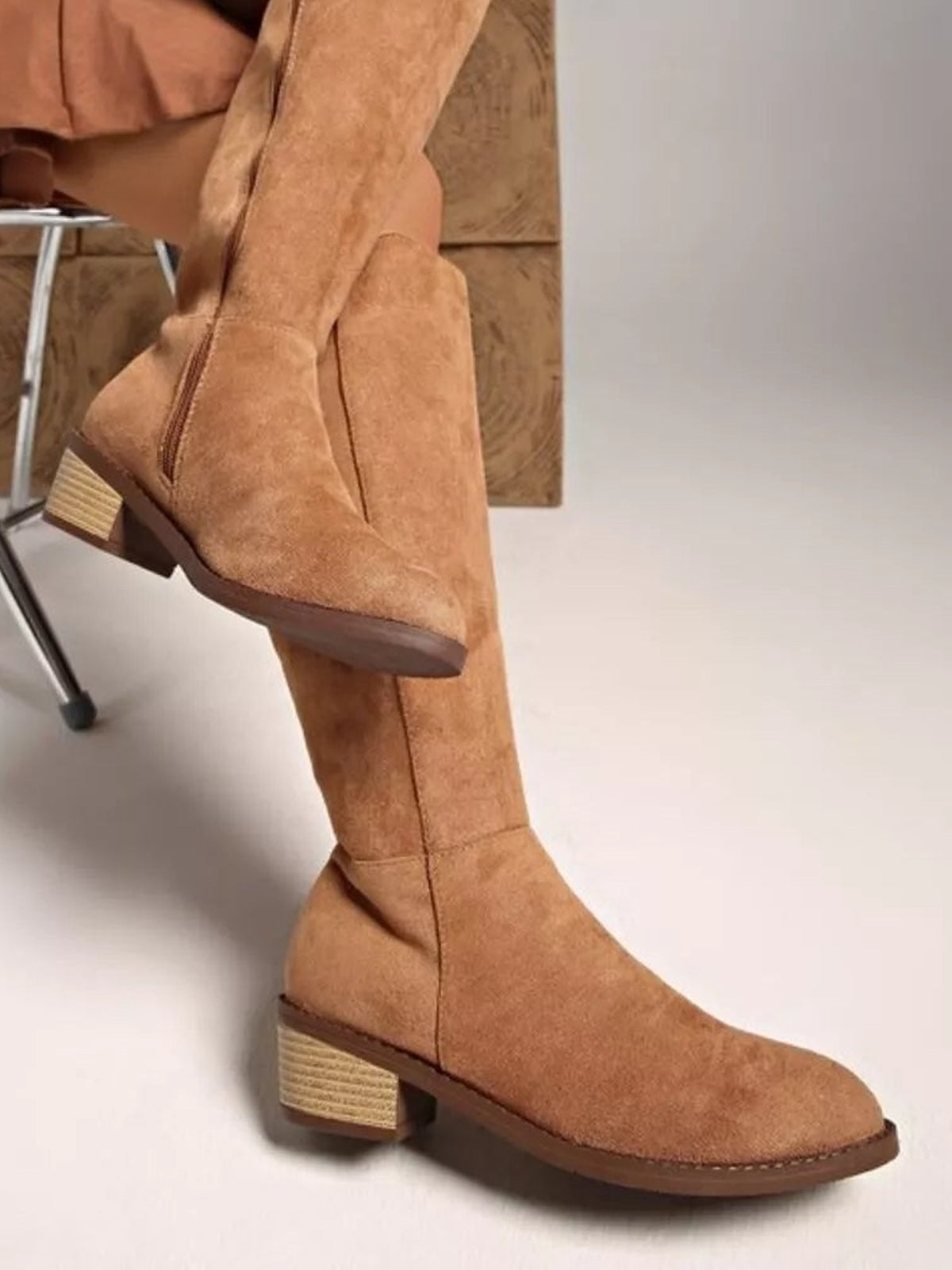 A seated model wears tan suede knee-high boots with slim shafts, side zippers, rounded toes, and sturdy mid-height block heels.