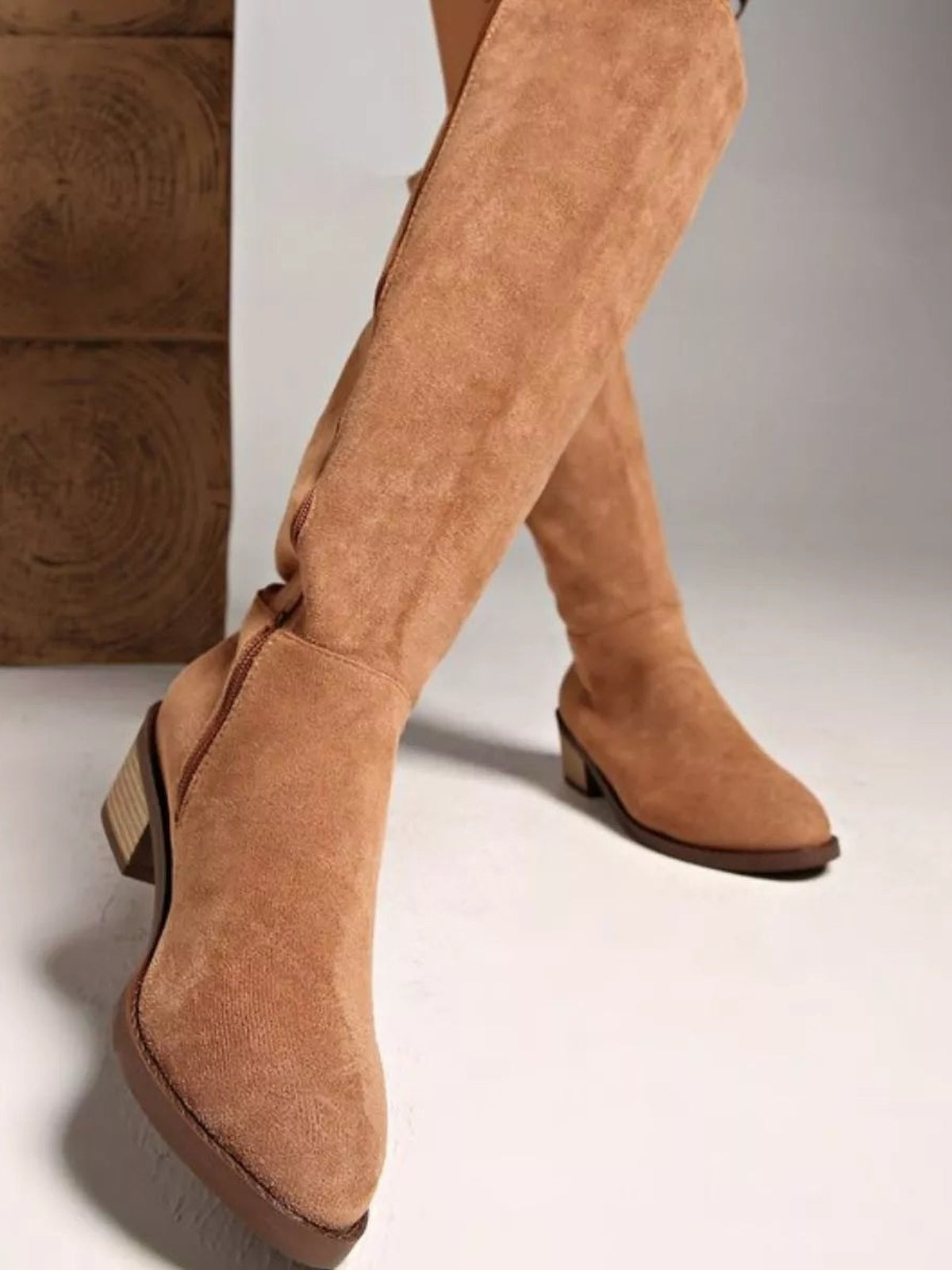 Tan suede knee-high boots on crossed legs, featuring a sleek silhouette, pointed toes, visible seam construction, and mid-height block heels with rubber soles.