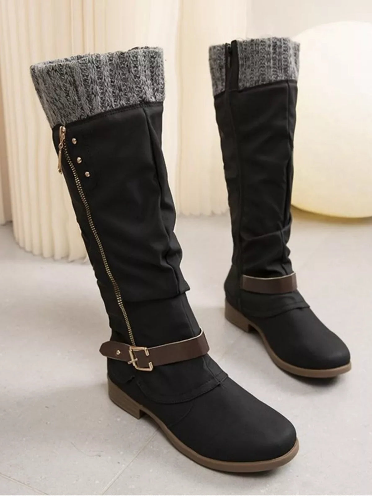 Black knee-high slouch boots with gray knit cuffs, low heels, side zippers, brown buckle straps, rounded toes, and rubber soles.